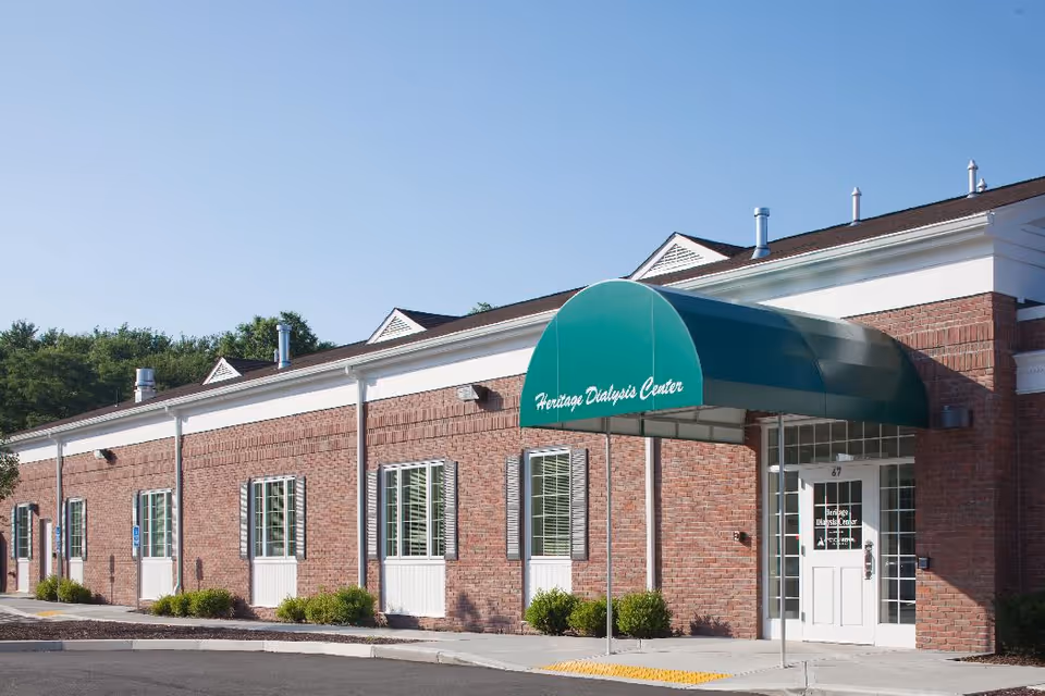 Exterior view of a single-story brick building with a green awning that reads 'Heritage Dialysis Center' over the entrance door. The building has several windows with white shutters and is surrounded by small bushes and a paved driveway.