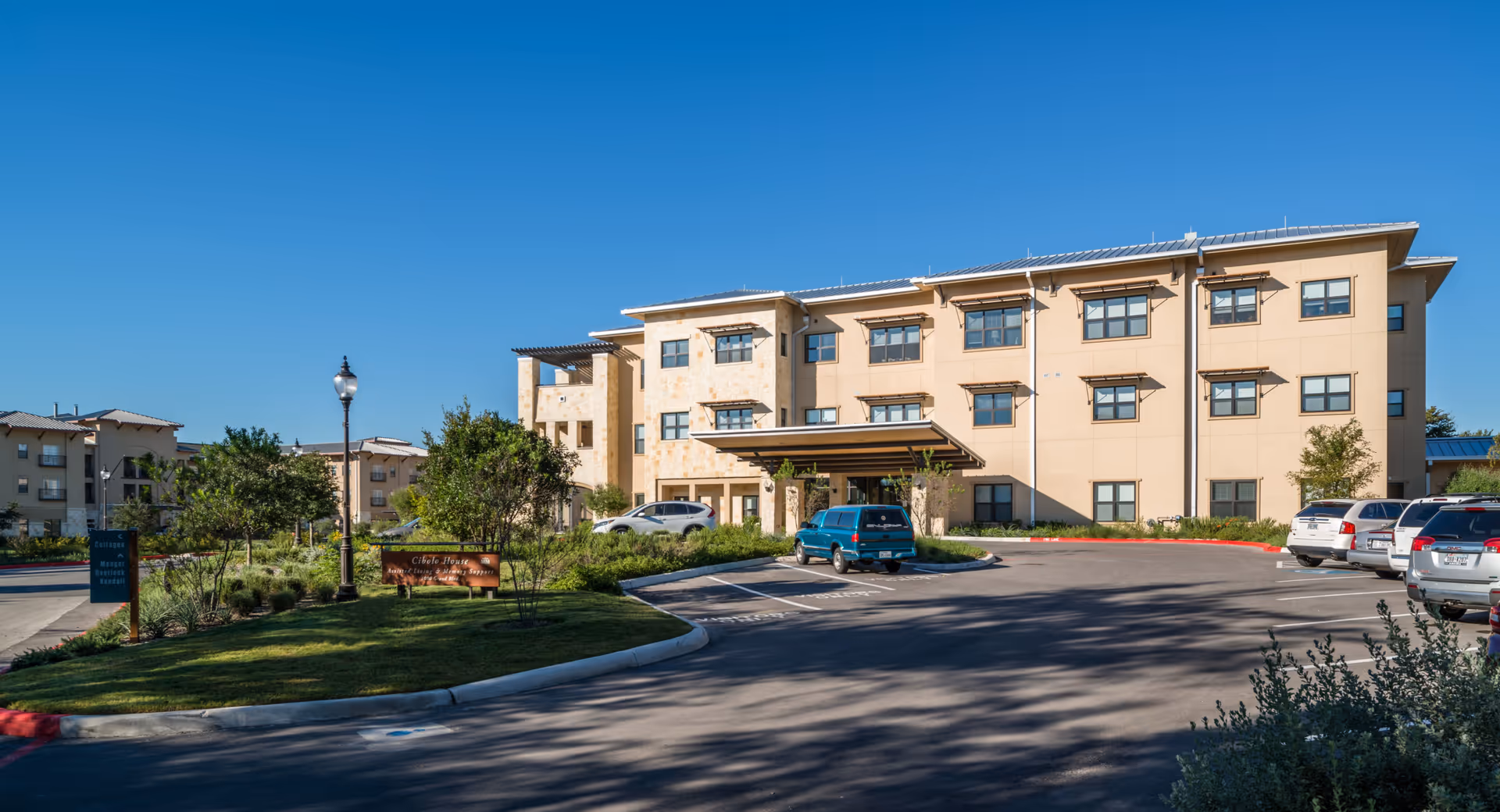 Three-story beige assisted living building with a covered entrance, parked cars, and landscaped grounds under a clear blue sky.