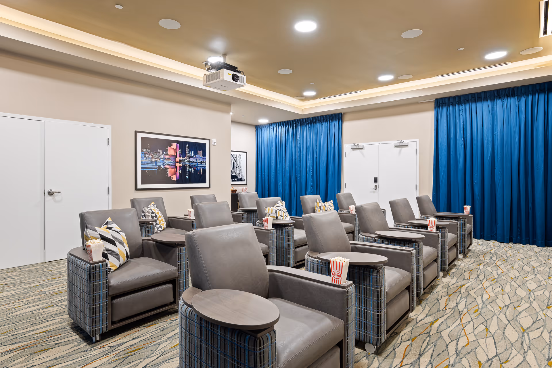 A small theater room with two rows of comfortable gray armchairs, each equipped with a small attached table. The chairs have patterned pillows and popcorn containers placed on the tables. The room has blue curtains along the back wall, a projector mounted on the ceiling, and framed artwork on the side wall. The carpet has a modern pattern with neutral tones.