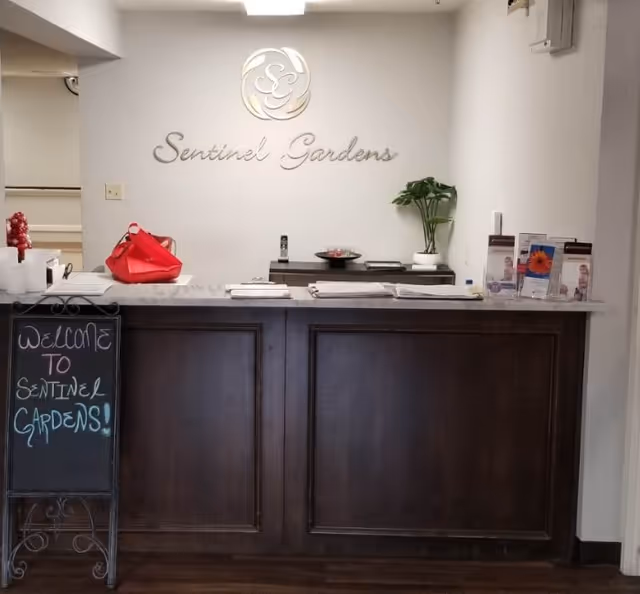 Reception desk area with a 'Sentinel Gardens' wall sign, brochures, a potted plant, and a chalkboard welcome sign.