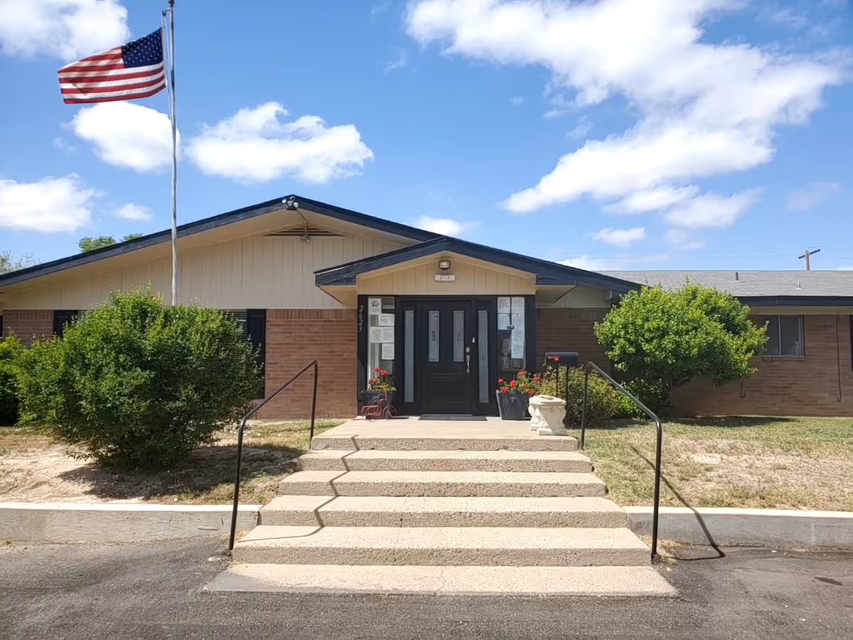 Front entrance of a single-story brick assisted living building with steps leading to double doors, potted flowers, shrubs, and an American flag.