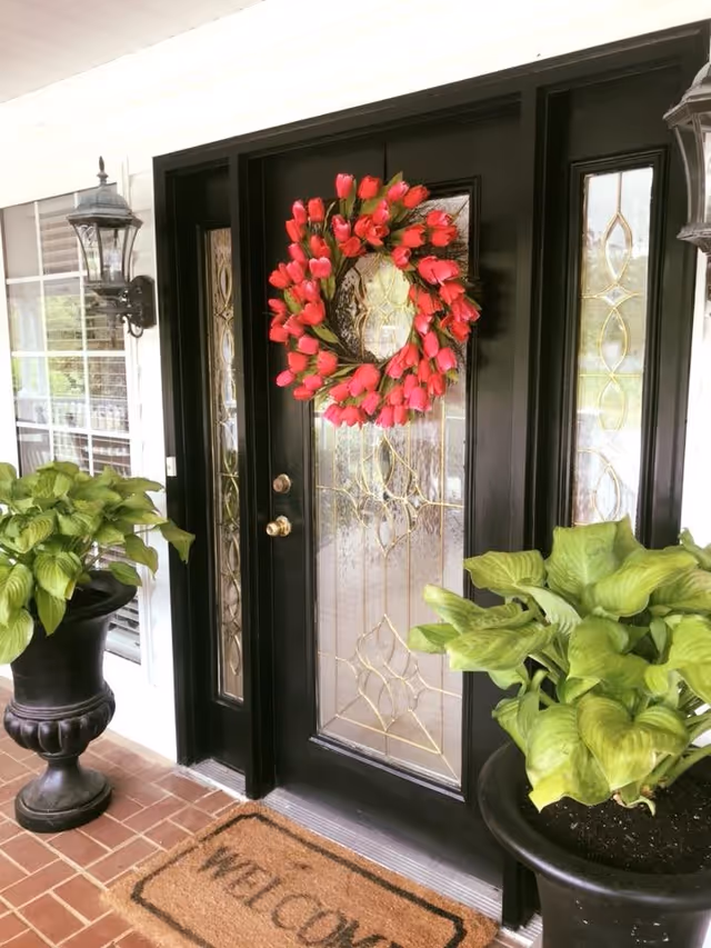 Front entrance door with decorative glass panels and a red tulip wreath hanging on it. Two large black planters with green leafy plants flank the door. A welcome mat is placed on the brick floor in front of the door. Two black wall-mounted lantern lights are on either side of the door.