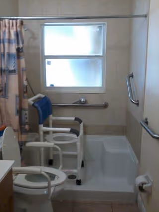 A bathroom with accessibility features including a raised toilet with armrests, a shower chair, grab bars on the walls, a bathtub with a built-in seat, a shower curtain, and a frosted window providing natural light.