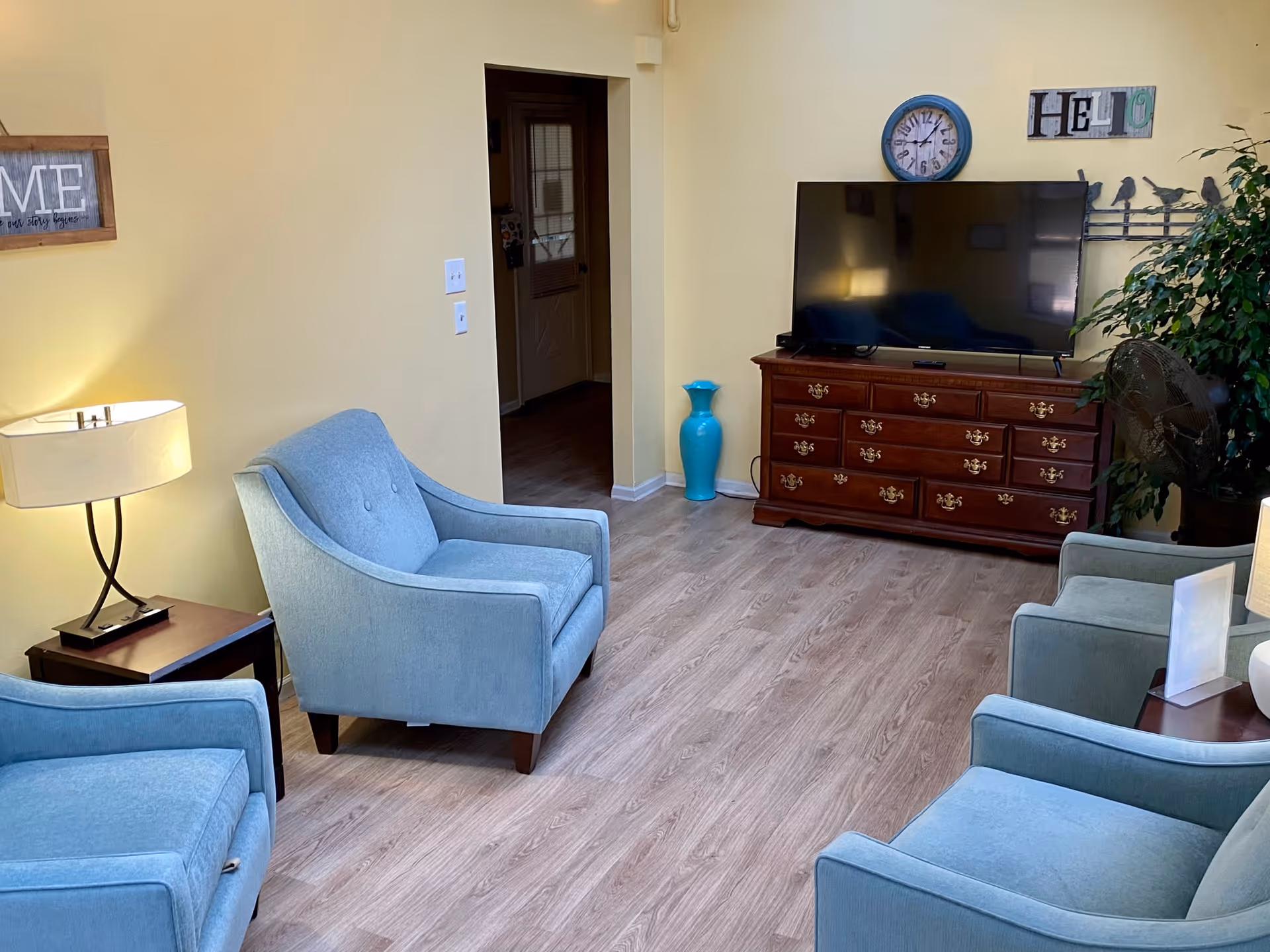 A cozy living room with four light blue armchairs arranged around a wooden TV stand holding a large flat-screen television. The walls are painted yellow and decorated with a clock, a 'HELLO' sign, and a framed 'HOME' sign. There is a floor lamp on a small wooden side table and a large green plant next to a standing fan.