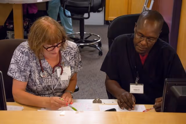 Two healthcare professionals sitting at a desk, reviewing and marking documents. One is a woman with glasses and a stethoscope around her neck, and the other is a man wearing glasses and a black scrub top. They appear focused on their work in an office setting.