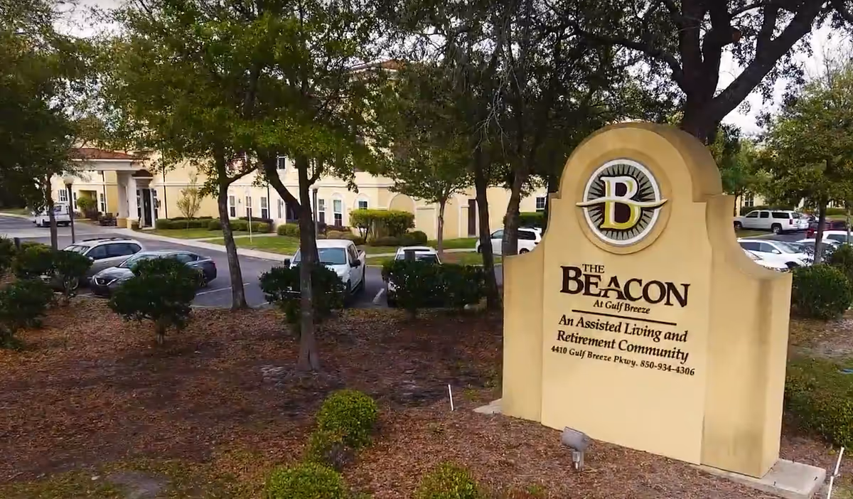 Outdoor view of The Beacon at Gulf Breeze assisted living and retirement community sign with the building and parking lot in the background surrounded by trees and landscaping.