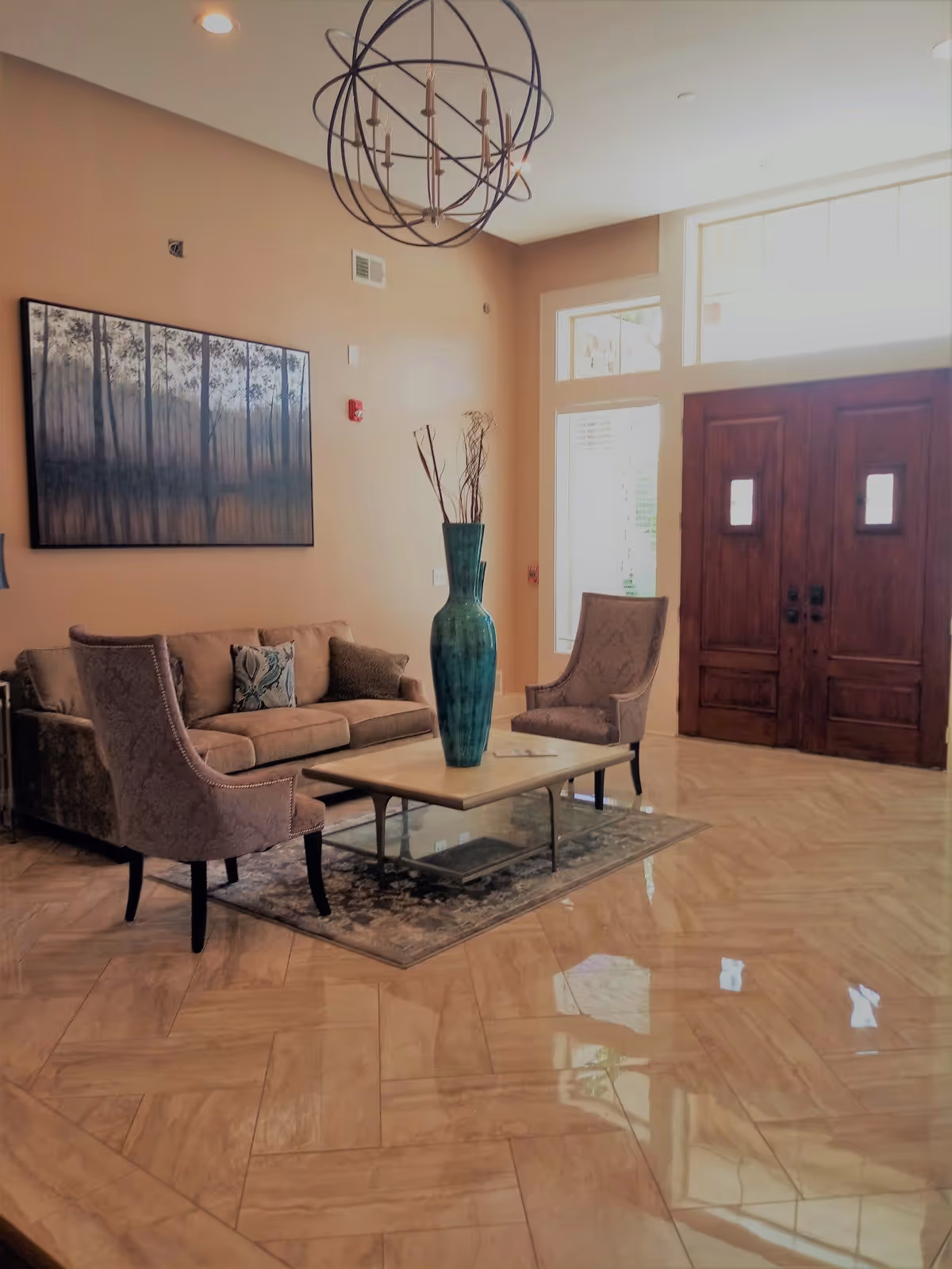 A cozy living room area with a beige sofa, two upholstered chairs, a rectangular coffee table with a tall green vase holding decorative branches, a large painting of a forest on the wall, and a modern spherical chandelier hanging from the ceiling. The room has large wooden double doors and windows letting in natural light, with a polished tile floor and a small area rug under the coffee table.