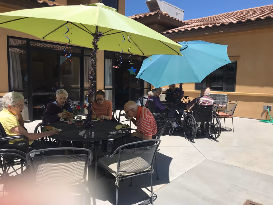 Several elderly people sitting in wheelchairs and regular chairs around two outdoor tables with large umbrellas, enjoying a meal in a sunny courtyard area of a senior living facility.