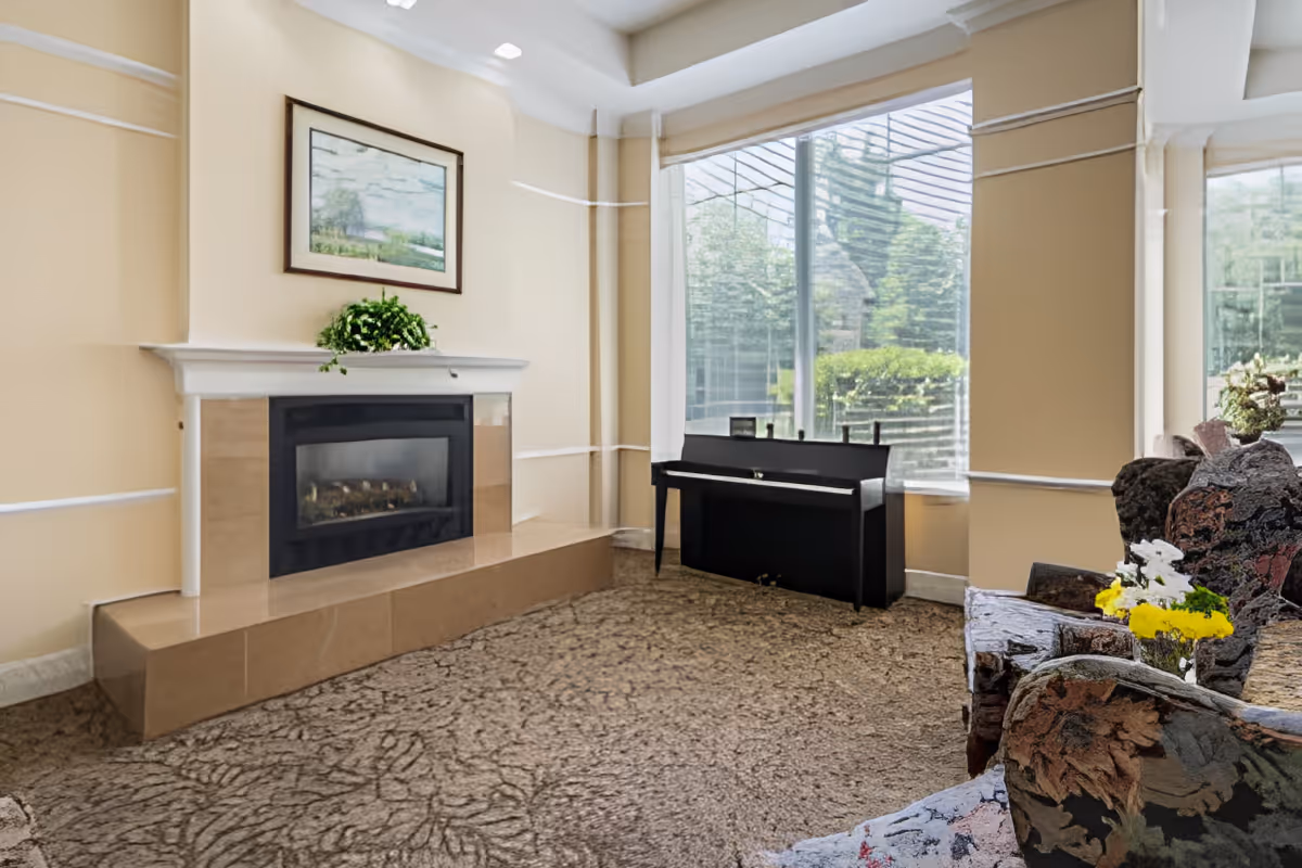 A cozy living room area with a beige carpet, a fireplace with a white mantel, a framed landscape painting above the fireplace, a black upright piano near large windows with a view of greenery outside, and floral-patterned armchairs with a small table holding a vase of yellow and white flowers.