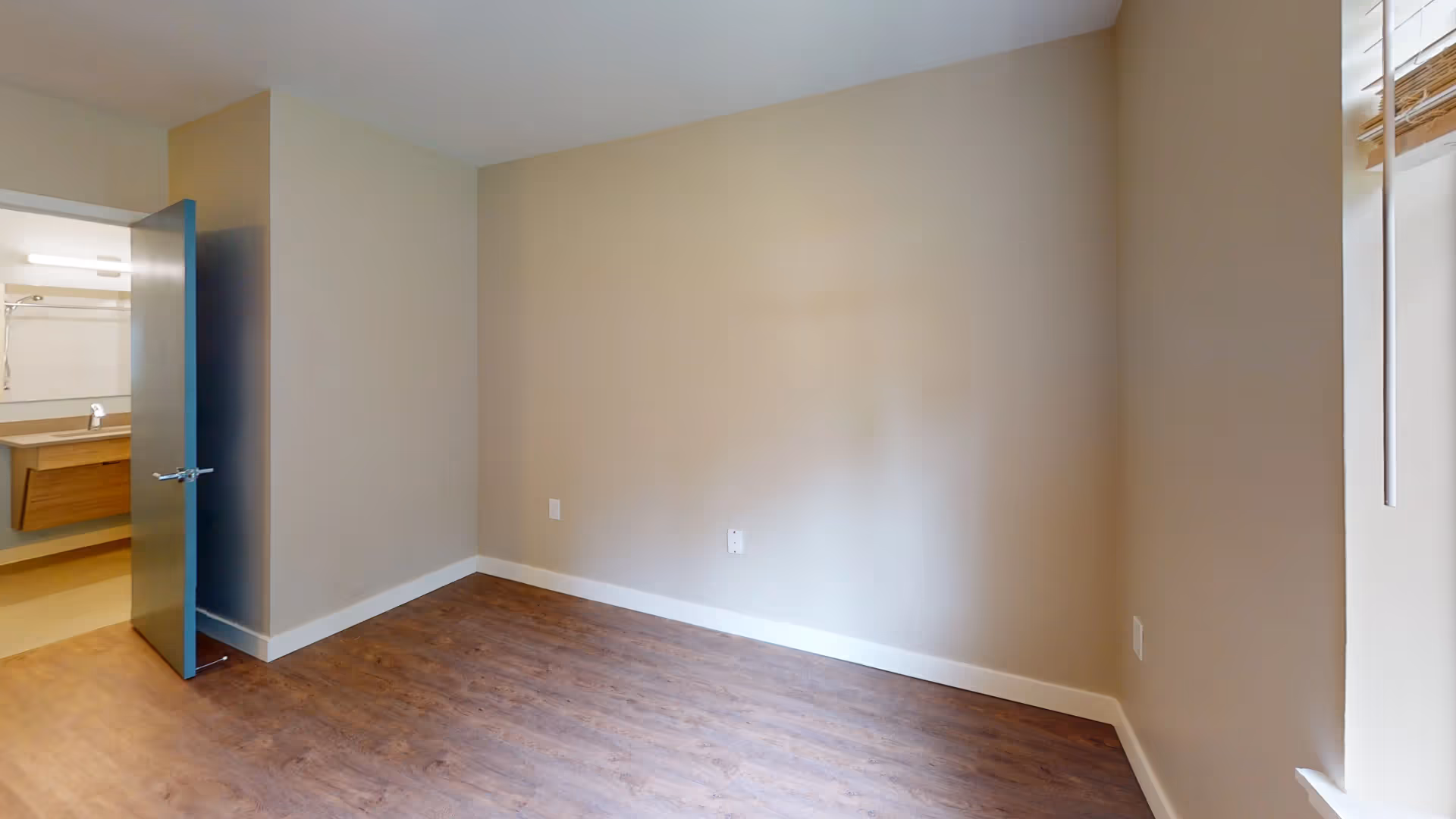 Empty small room with wood-look flooring, beige walls, a window on the right, and an open door revealing a sink/vanity.