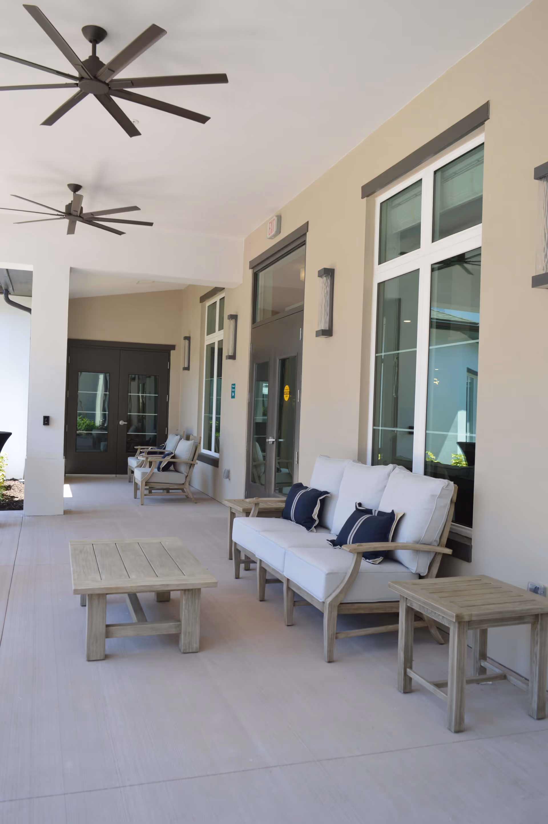 Covered outdoor patio area with beige walls, large windows, and ceiling fans. The patio is furnished with cushioned wooden seating including a sofa with white cushions and blue pillows, armchairs, and wooden tables.