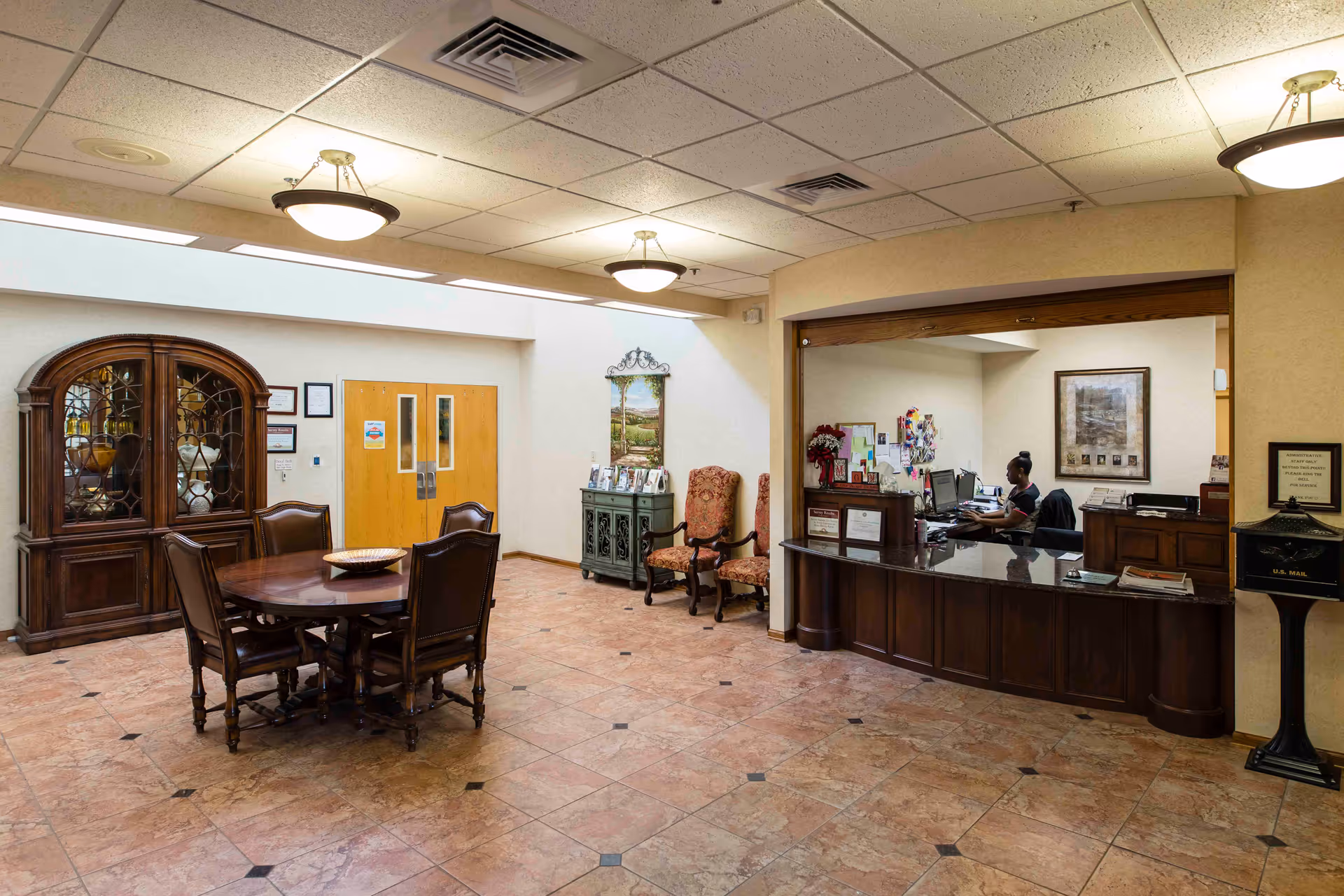 Interior view of a reception area in a senior living facility with a wooden reception desk where a staff member is working on a computer. The room has tiled flooring, a round wooden table with four chairs, a wooden cabinet with glass doors, two upholstered chairs, and framed artwork on the walls. Ceiling lights provide illumination.