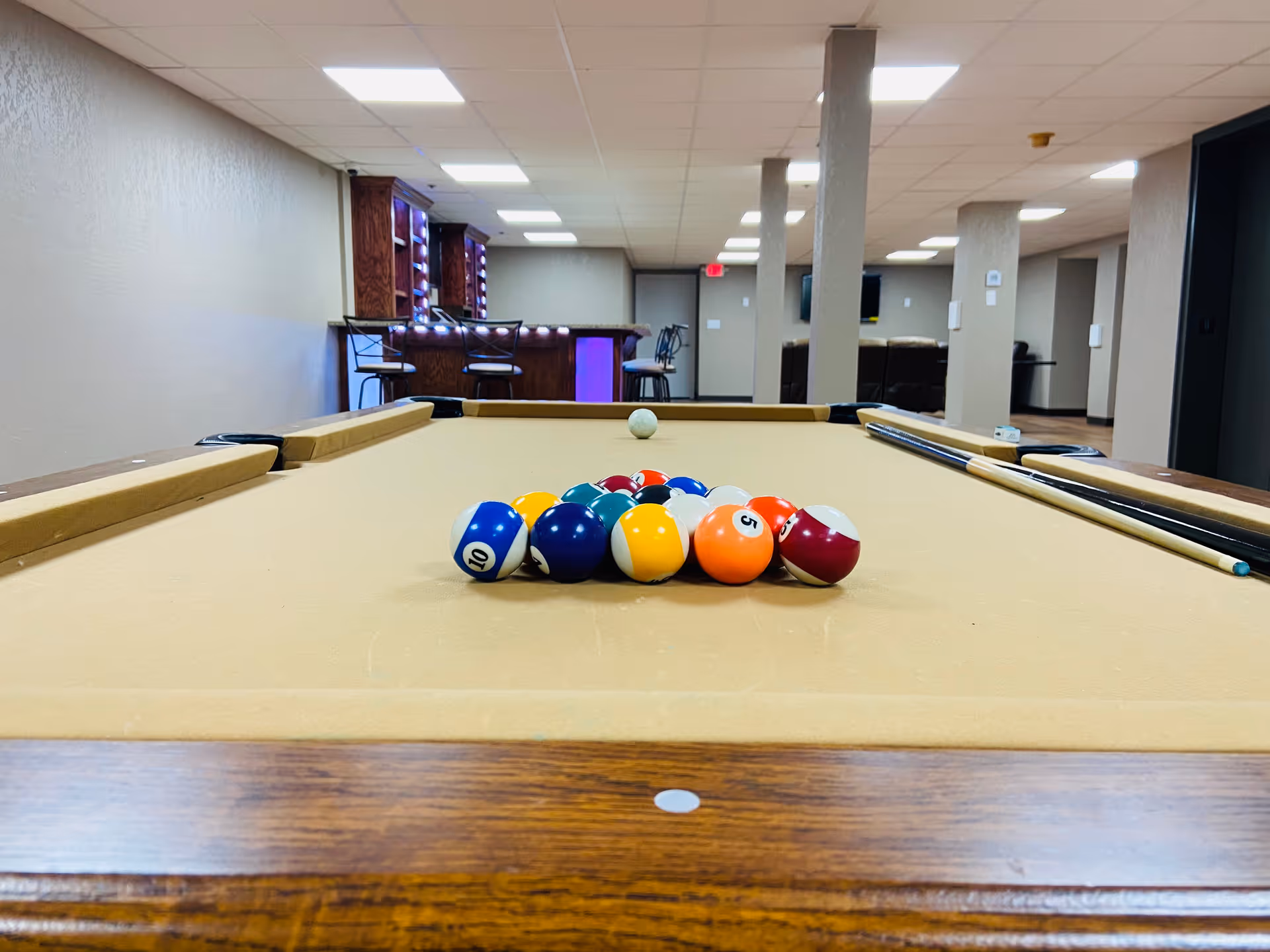 Racked pool balls on a billiards table in a recreation room with a bar and seating in the background.
