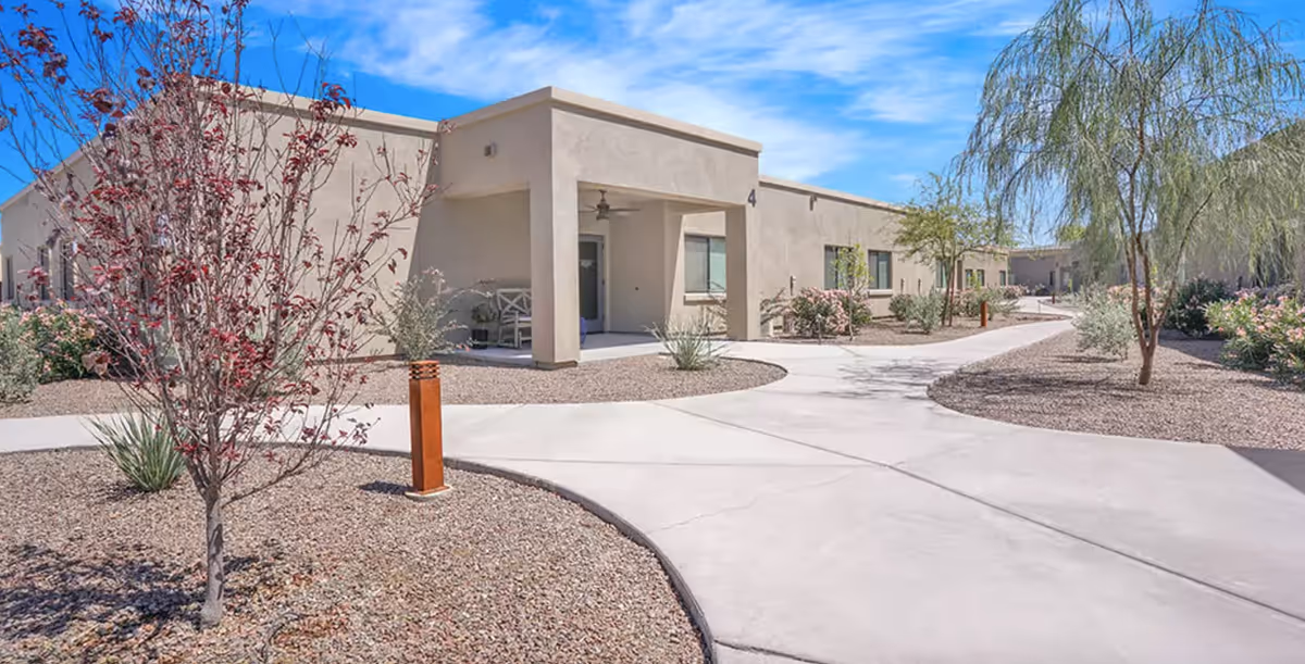 Single-story beige building with a covered entry and curved concrete walkways through desert landscaping under a blue sky.