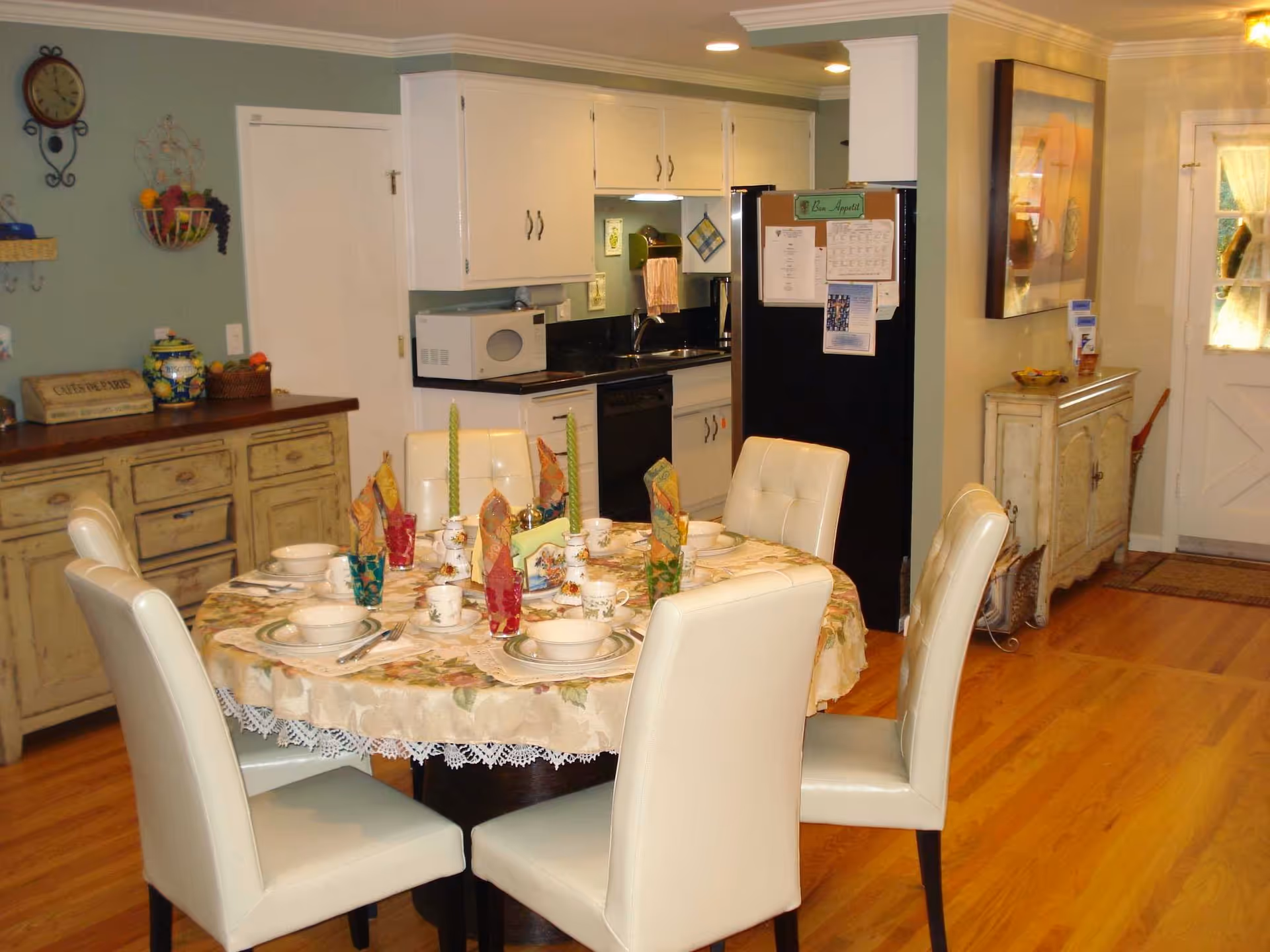A cozy dining area with a round table set for six people, featuring floral tablecloth, colorful napkins, cups, and plates. The room has light-colored leather chairs, wooden flooring, and a kitchen area in the background with white cabinets, a black refrigerator, microwave, and sink. There are decorative items on the walls and furniture, including a clock and a basket of fruit.