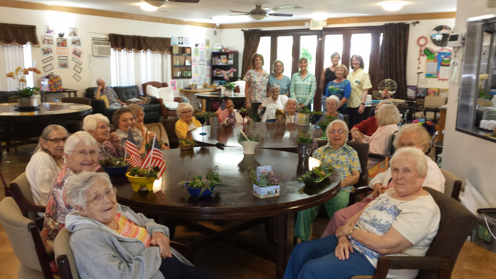 A group of elderly residents and staff members gathered around a large, oval wooden table in a common room. The table is decorated with small potted plants and miniature American flags. The room has comfortable seating, shelves with books and games, and large windows with curtains letting in natural light.
