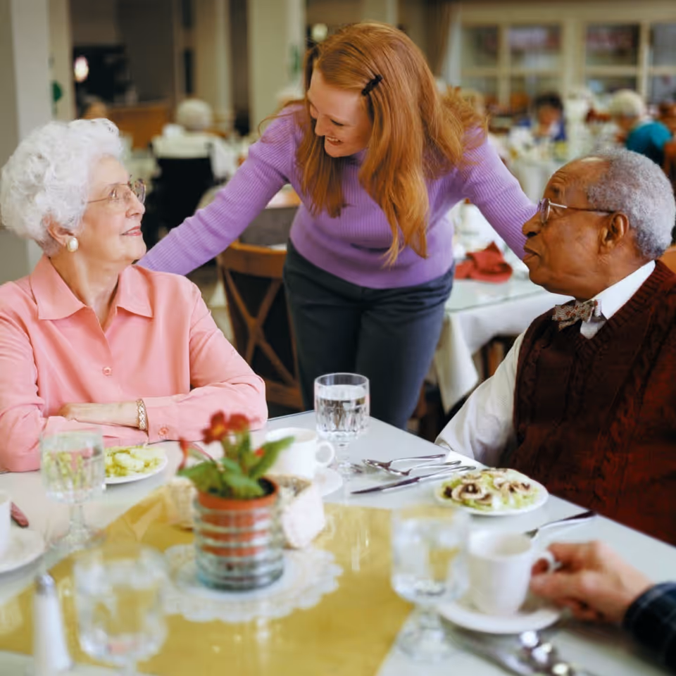 A caregiver leans and smiles while talking with two elderly residents seated at a dining table in a communal dining room.