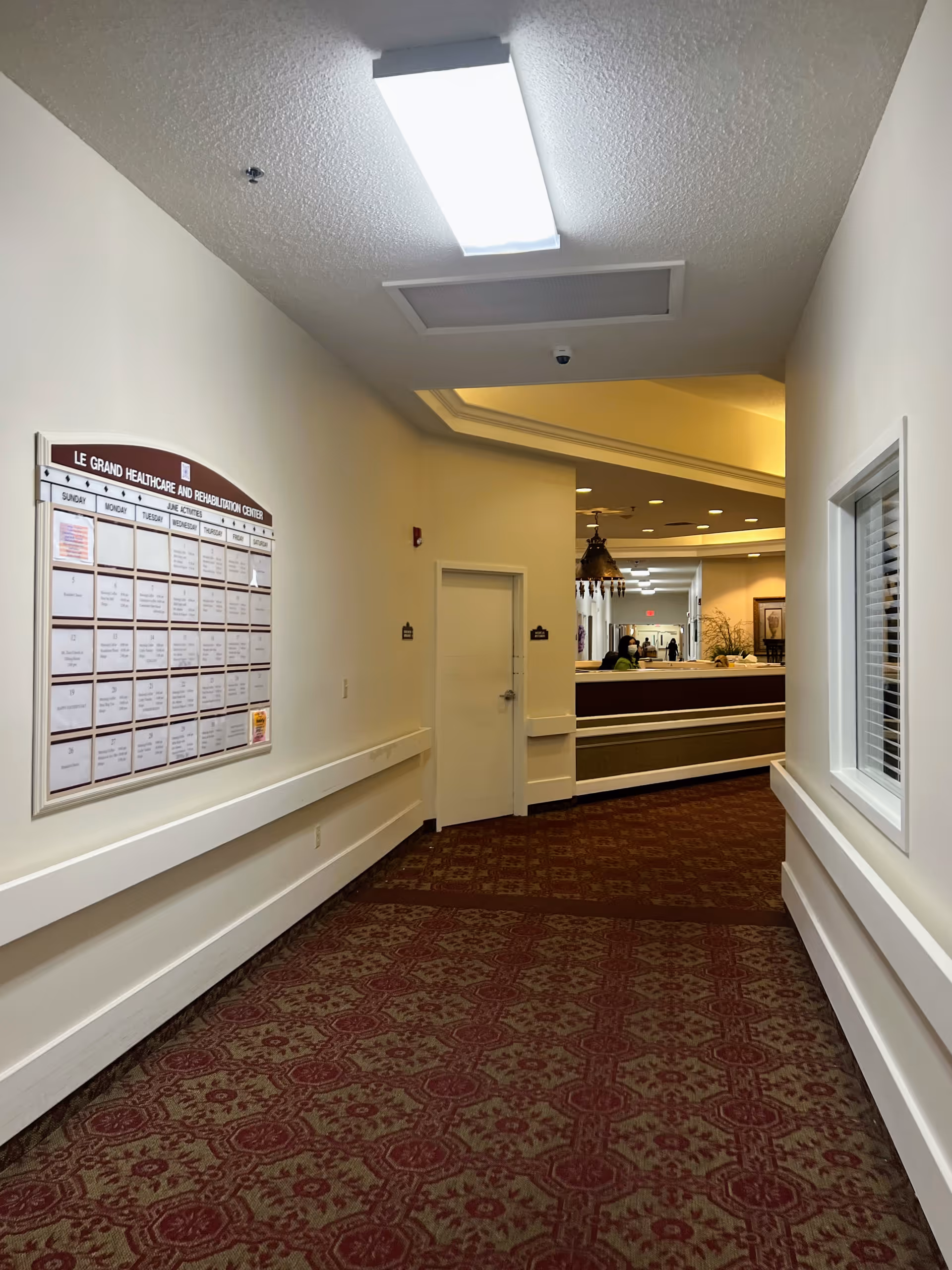 Carpeted hallway in a healthcare facility with a bulletin board on the left leading to a reception desk area.