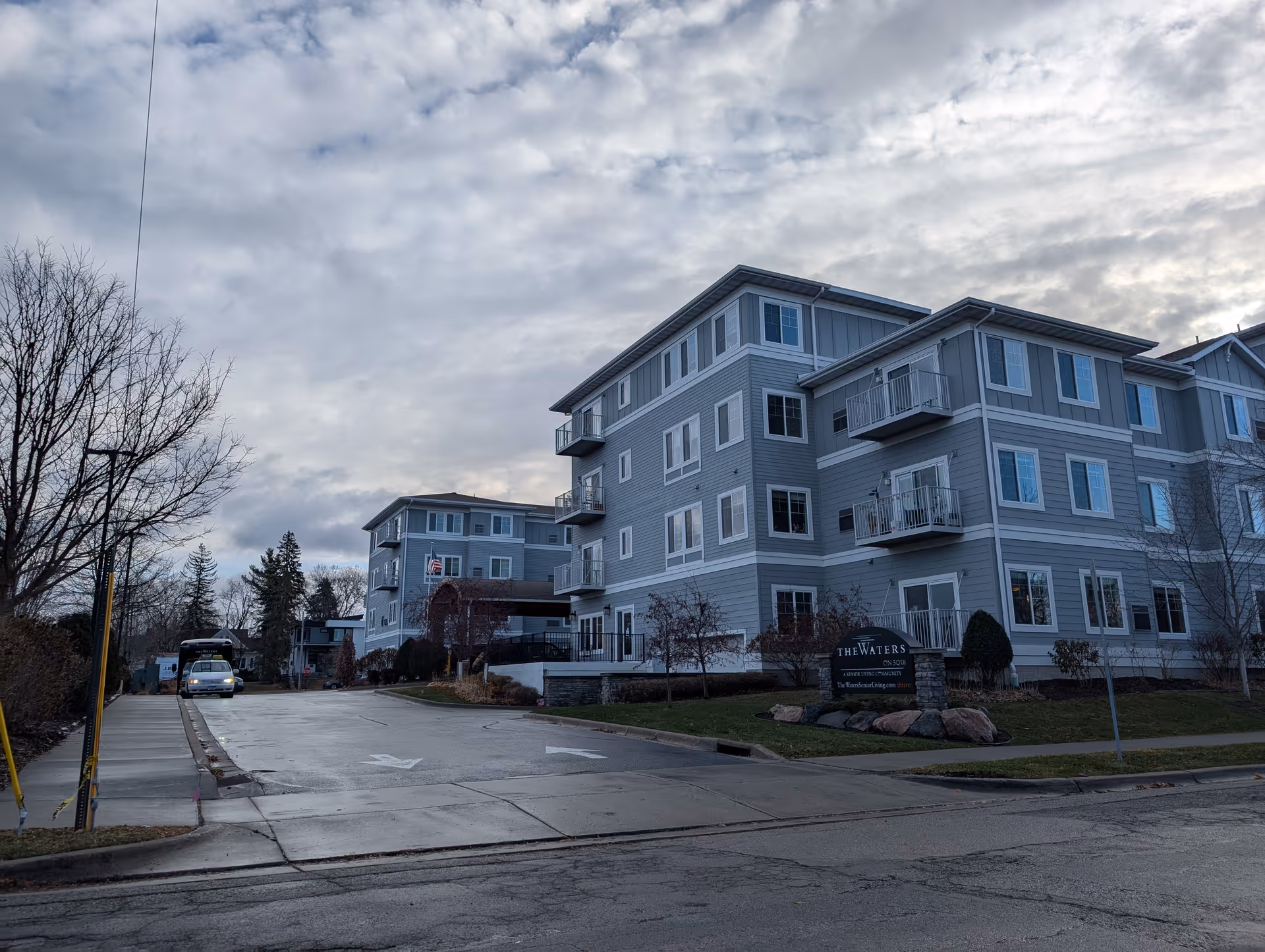 Front exterior of a multi-story gray senior living building labeled 'The Waters' with a driveway and a vehicle parked to the left on a cloudy day.