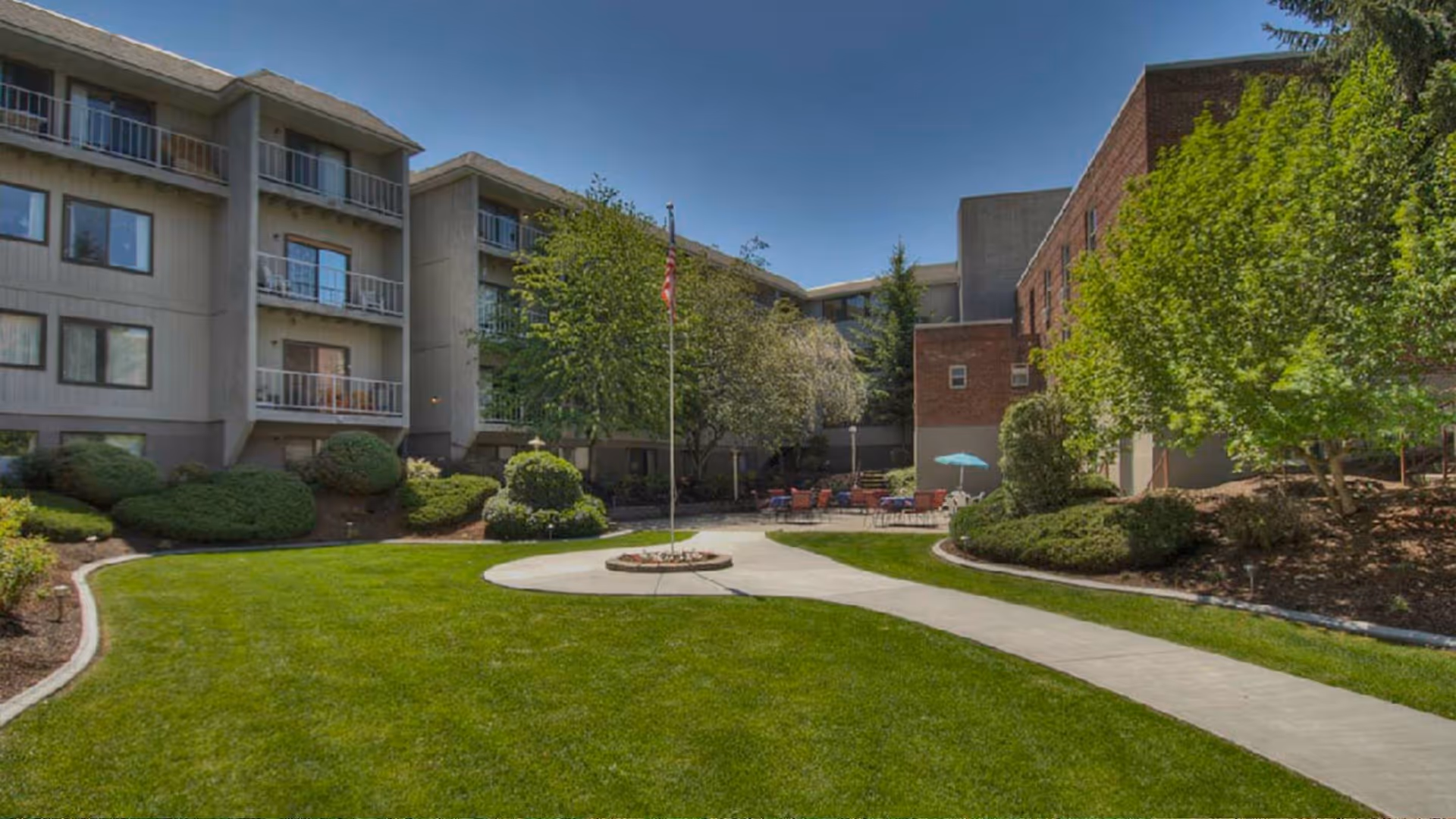 Outdoor courtyard area of a senior living facility with well-maintained green lawn, bushes, trees, a flagpole with an American flag, and a paved walkway. The courtyard is surrounded by multi-story buildings with balconies and windows. There are outdoor seating arrangements with chairs and tables under an umbrella.