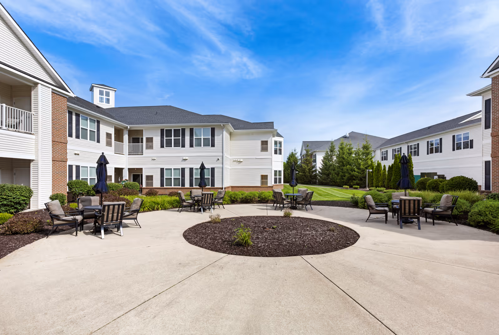 Courtyard with a circular plaza and outdoor seating surrounded by two-story senior living buildings under a blue sky.