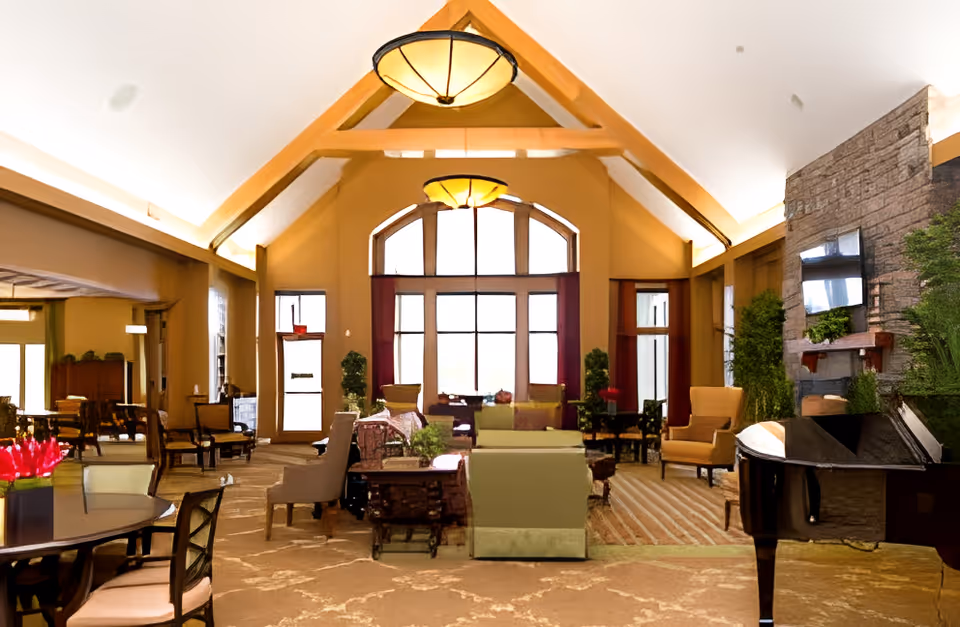 A spacious communal living room with a vaulted ceiling, large arched windows, multiple seating areas, a fireplace and a grand piano.