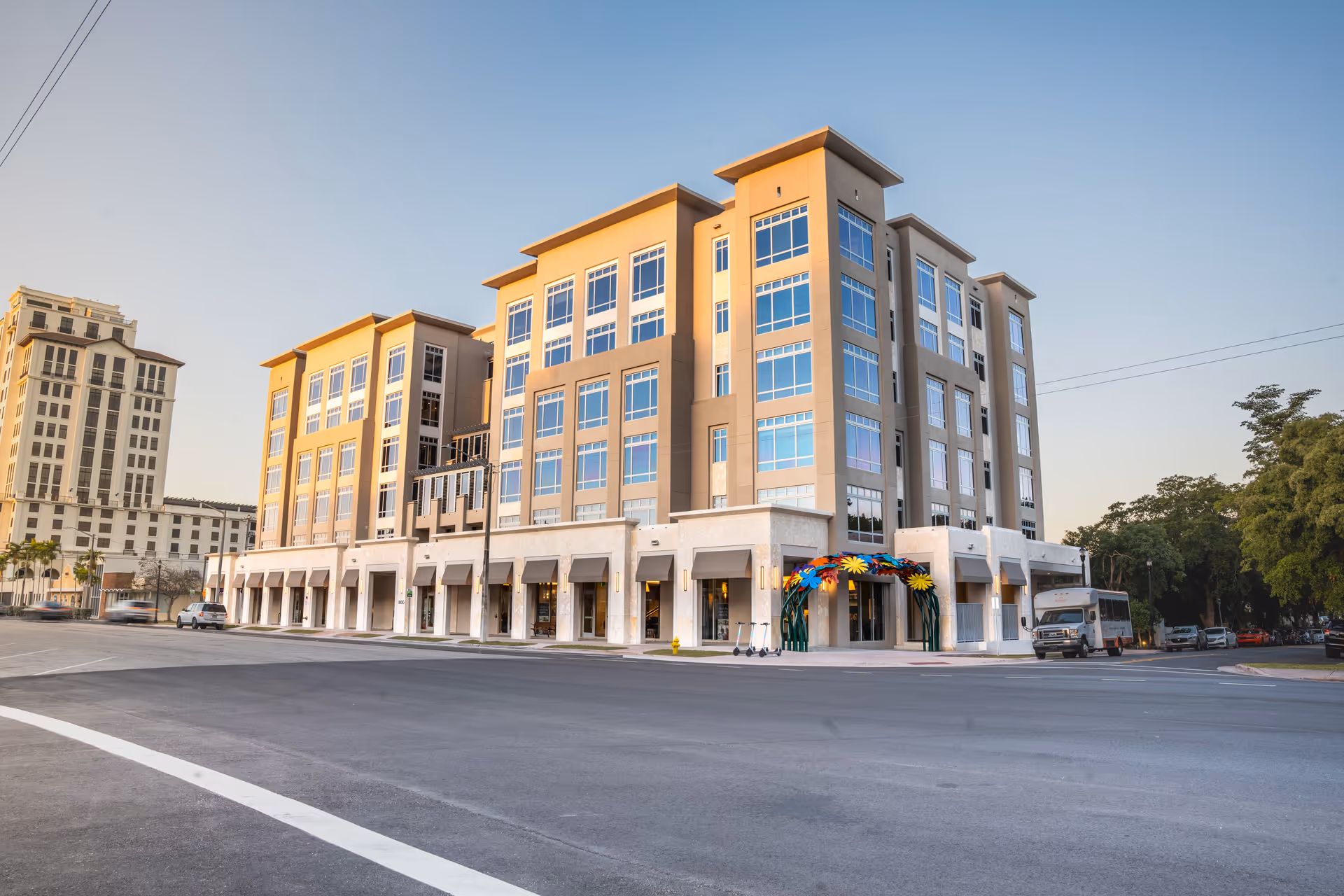 A multi-story modern building with large windows and a decorative entrance featuring colorful flower sculptures, situated on a street corner under a clear sky.