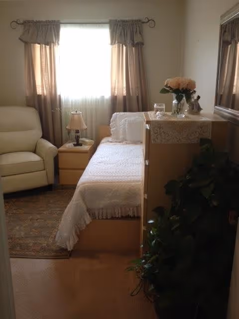 A cozy bedroom with a single bed covered in a white quilt, a wooden nightstand with a lamp and a book, a beige armchair, a window with sheer and brown curtains, a dresser with a lace runner, a vase of flowers, and a large green plant in the corner.