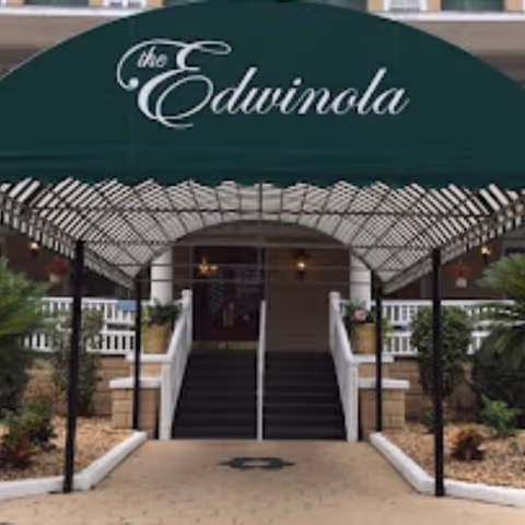 Entrance to a building with a green canopy that reads 'the Edwinola' above a set of stairs flanked by white railings and potted plants.