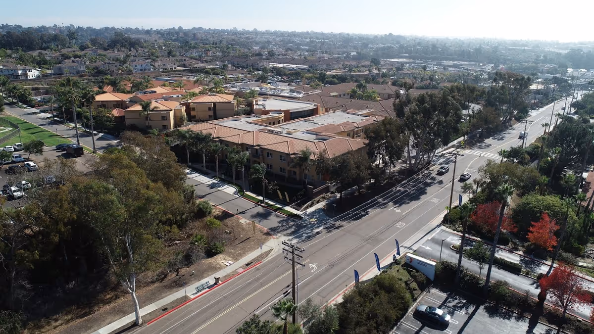 Aerial view of a residential area featuring a large retirement community with multiple buildings with red-tiled roofs, surrounded by trees and streets. The image shows a main road with cars and sidewalks, and a mix of greenery and urban development extending into the distance under a clear sky.