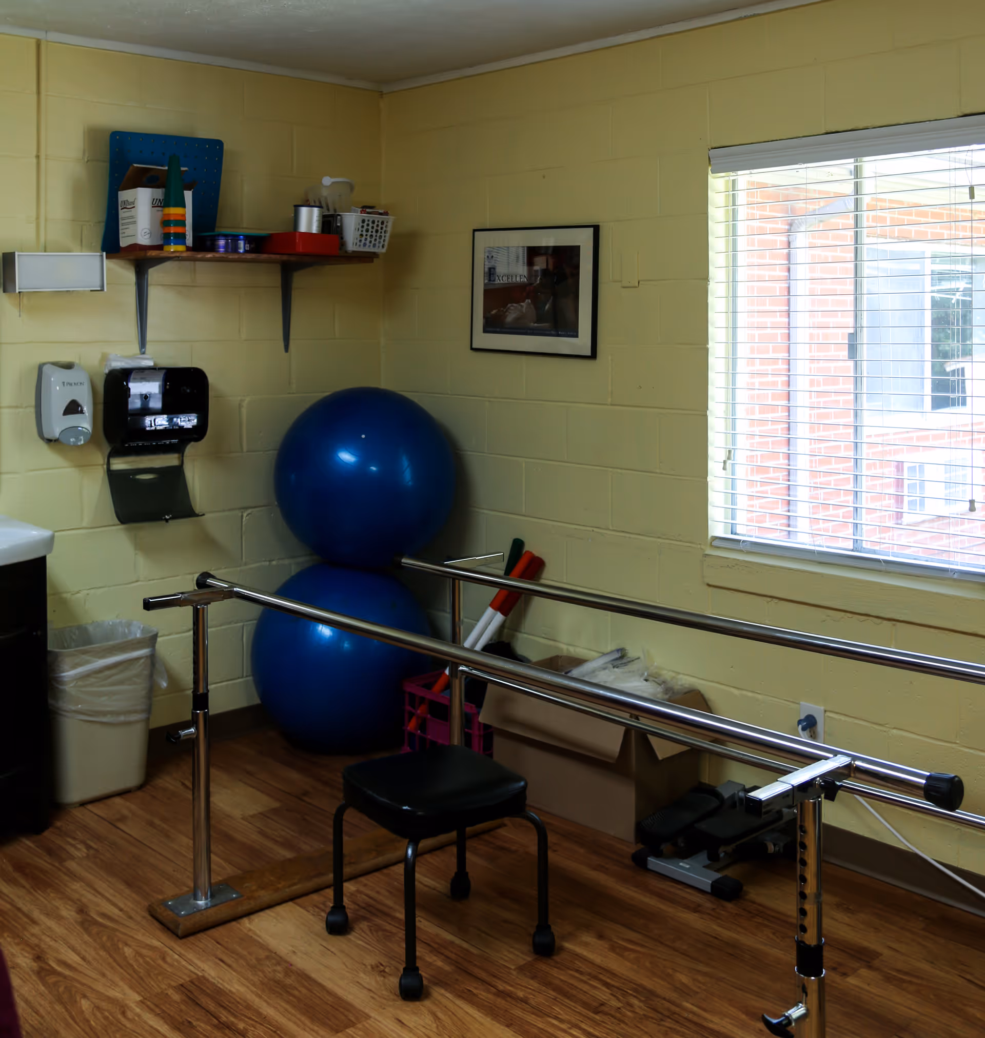 A small rehabilitation room with two large blue exercise balls stacked against a yellow brick wall, parallel bars for walking support, a black stool with wheels, a window with blinds letting in natural light, a shelf with various items, a soap dispenser, paper towel dispenser, and a trash can.
