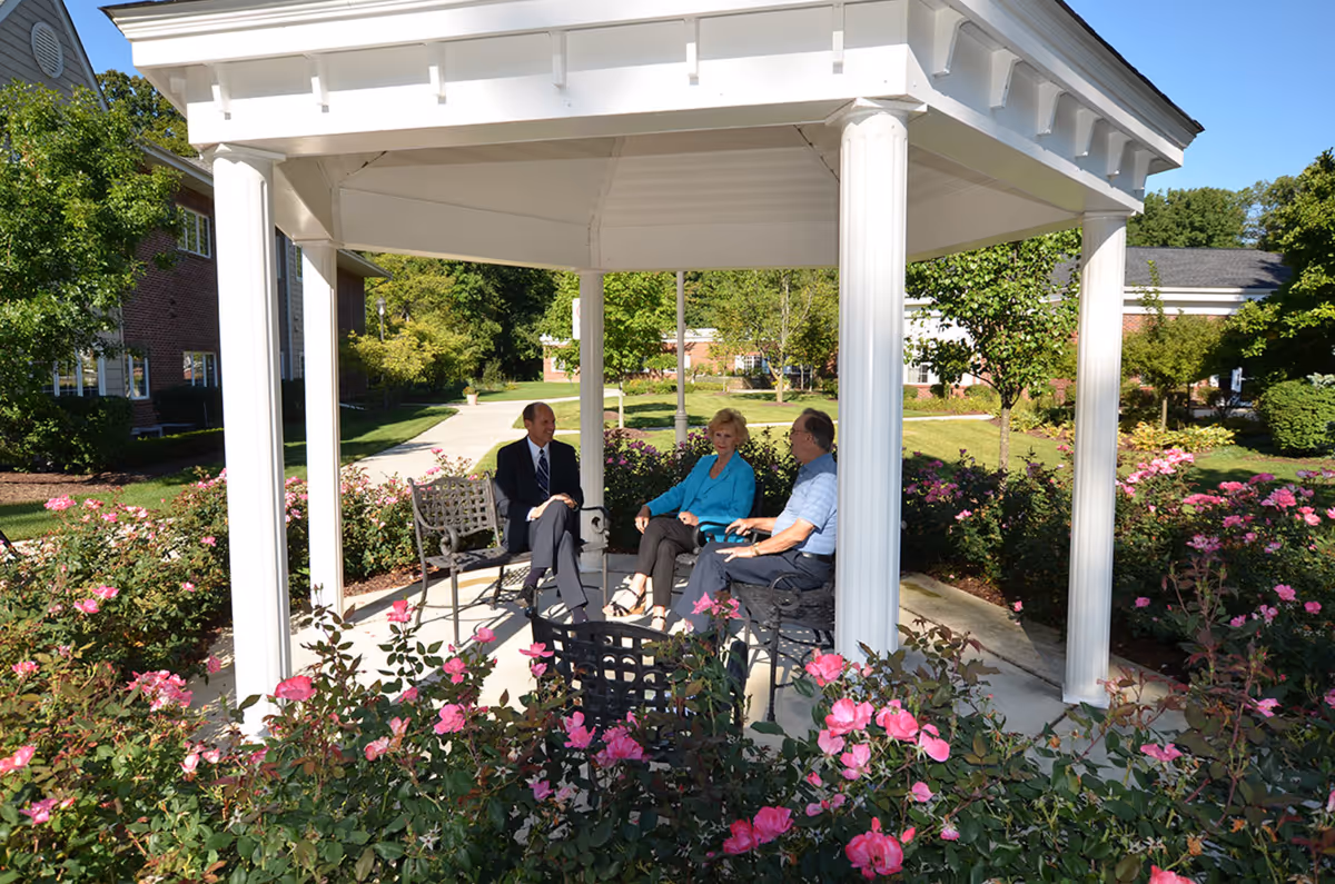 Three elderly people sitting and conversing under a white gazebo surrounded by blooming pink flowers and greenery in an outdoor garden area of a retirement community.