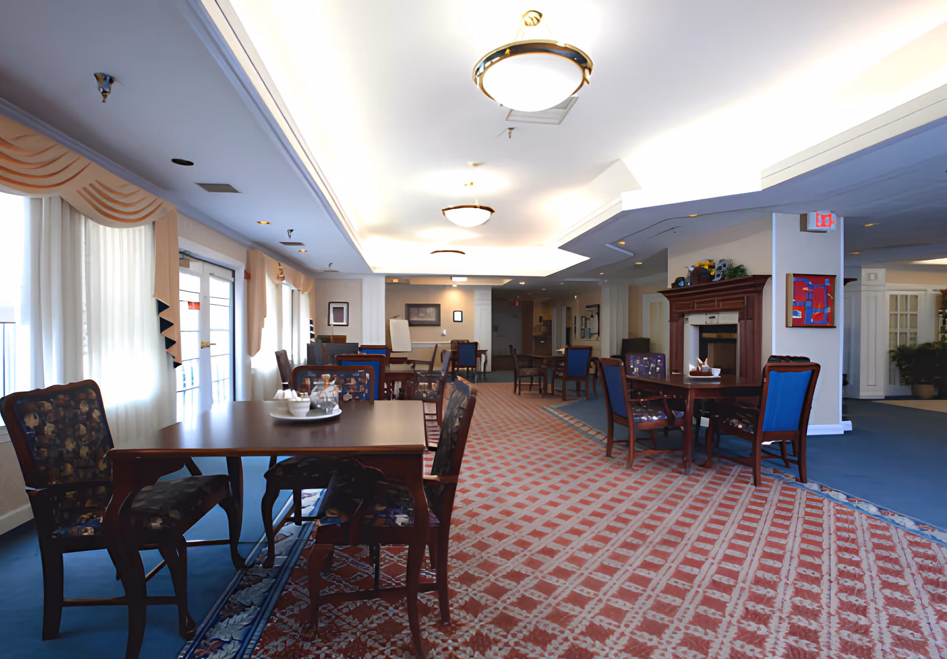 Spacious senior living community dining/lounge area with tables and chairs, patterned carpet, and a fireplace.