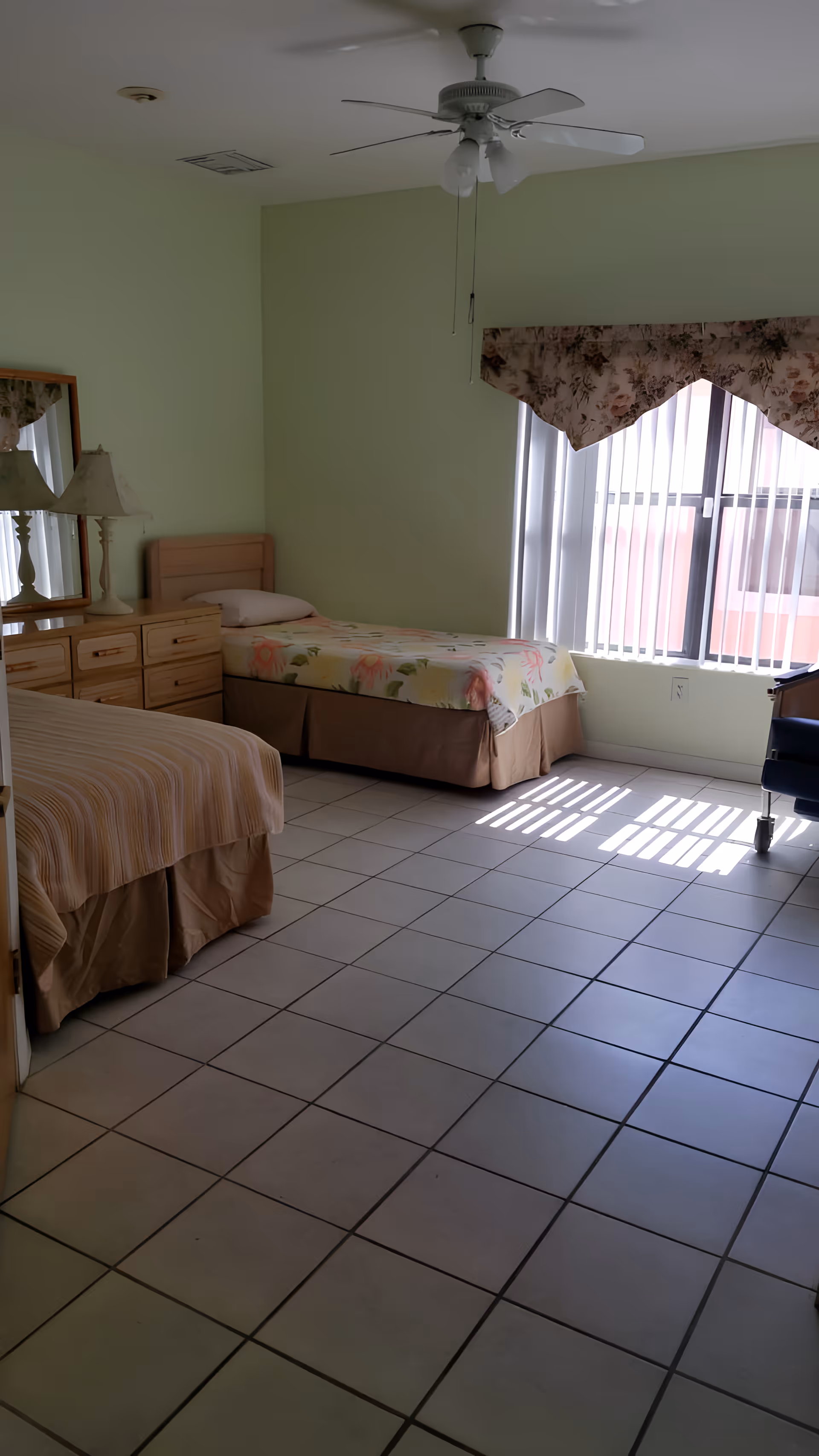 A bedroom in an assisted living facility with two single beds, one with a floral bedspread and the other with a striped bedspread. There is a wooden dresser with a mirror and a lamp on top. The room has light green walls, a ceiling fan with lights, and a window with vertical blinds and floral valance. The floor is tiled, and there is a wheelchair partially visible near the window.