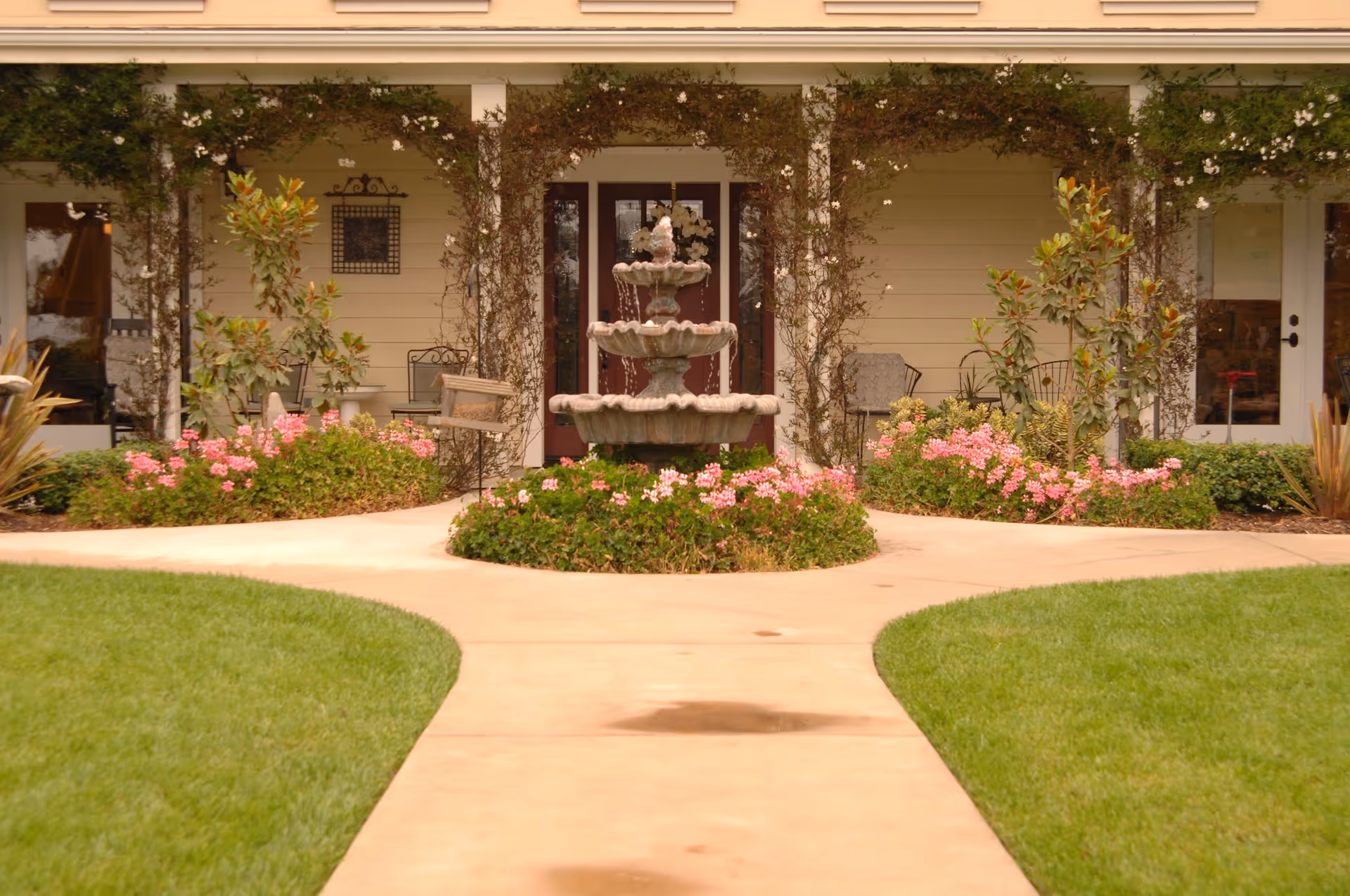 A landscaped outdoor area with a concrete pathway leading to a three-tiered water fountain surrounded by pink flowers and greenery. The background shows a building facade with glass doors and windows, some outdoor chairs, and climbing plants on the walls.