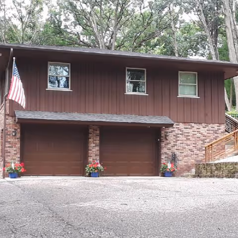 Exterior view of a two-story building with brown wooden siding and brick lower walls. The building has two closed brown garage doors, three windows on the upper floor, and an American flag mounted on the left side. There are potted plants with red flowers placed near the garage doors. Trees are visible in the background.