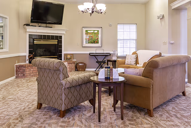 A cozy living room area with a patterned armchair and a brown sofa arranged around a small round wooden table. The room features a fireplace with a mounted flat-screen TV above it, a window with blinds, a framed picture on the wall, and a chandelier hanging from the ceiling. The carpet has a geometric pattern and the walls are painted in neutral tones.