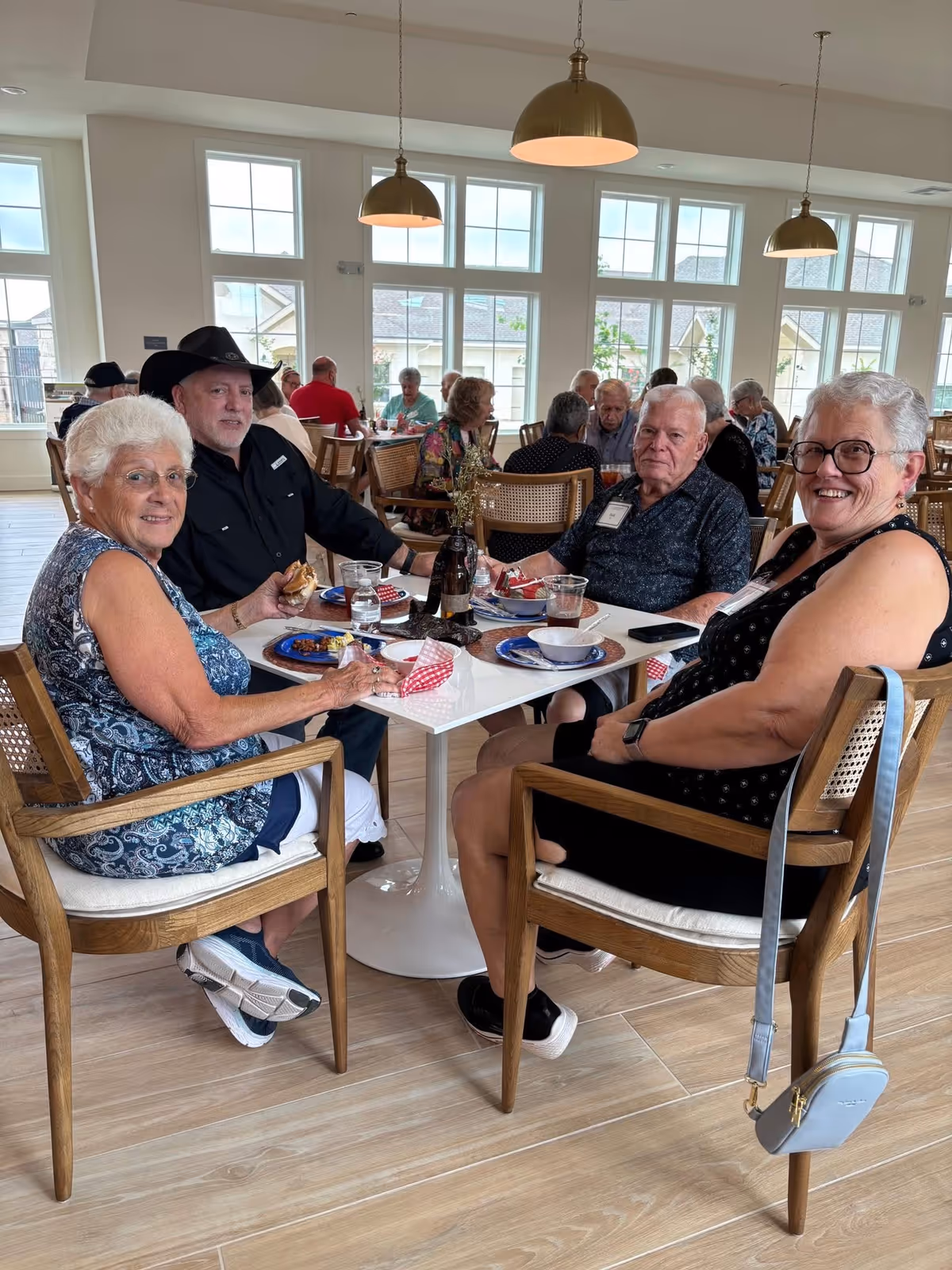 Four elderly people sitting around a square white table in a bright dining area with large windows and wooden chairs. They are enjoying a meal and smiling at the camera. Other seniors are seated at tables in the background. The room has wooden flooring and pendant lights hanging from the ceiling.