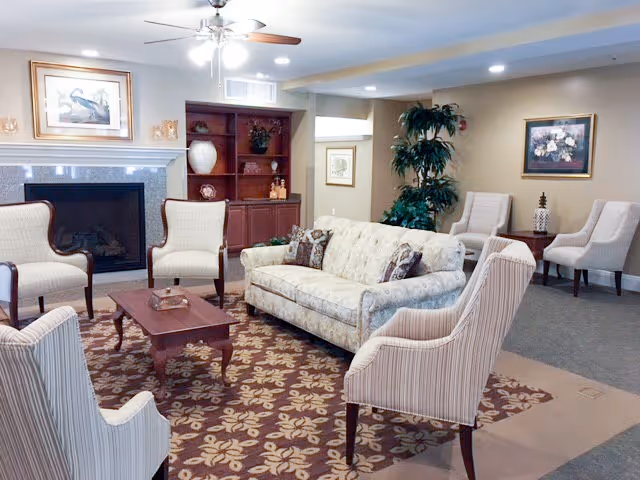 A cozy living room area in an assisted living facility featuring a floral patterned carpet, a beige sofa with decorative pillows, four striped armchairs arranged around a wooden coffee table, a fireplace with a framed picture above it, built-in wooden shelves with decorative items, a ceiling fan with lights, and a potted plant in the corner.