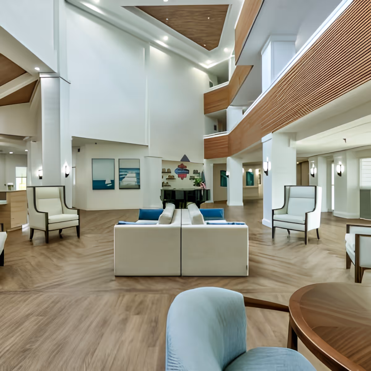 Spacious and modern senior living facility common area with high ceilings, wooden flooring, white walls, and contemporary furniture including armchairs and sofas arranged around a black grand piano. The space features wooden balcony railings on the upper floors and wall-mounted lights.