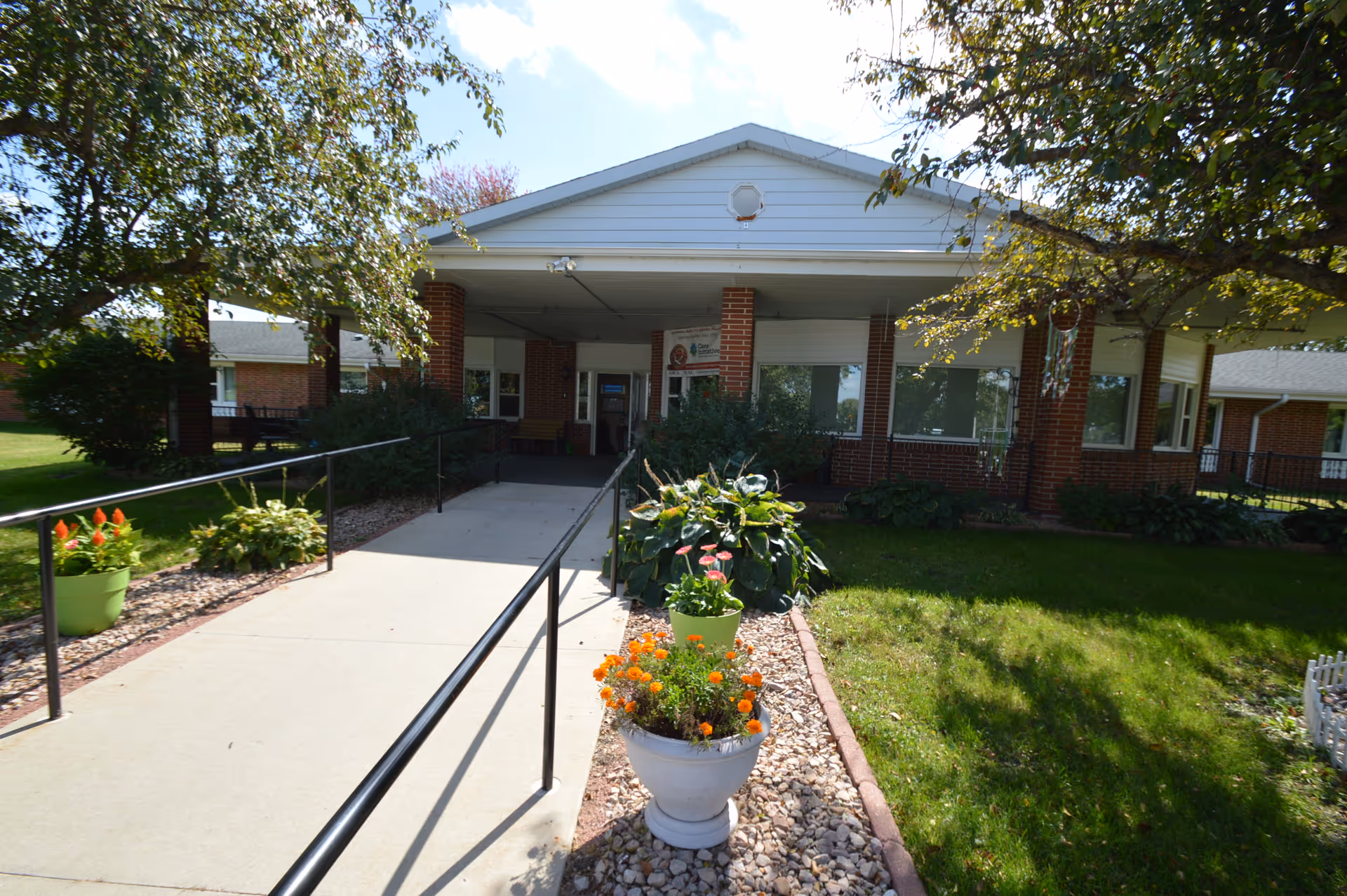 Front exterior view of a single-story brick building with a covered entrance, a concrete walkway with black handrails, and flower pots with colorful flowers on either side. Trees and green grass surround the entrance area.