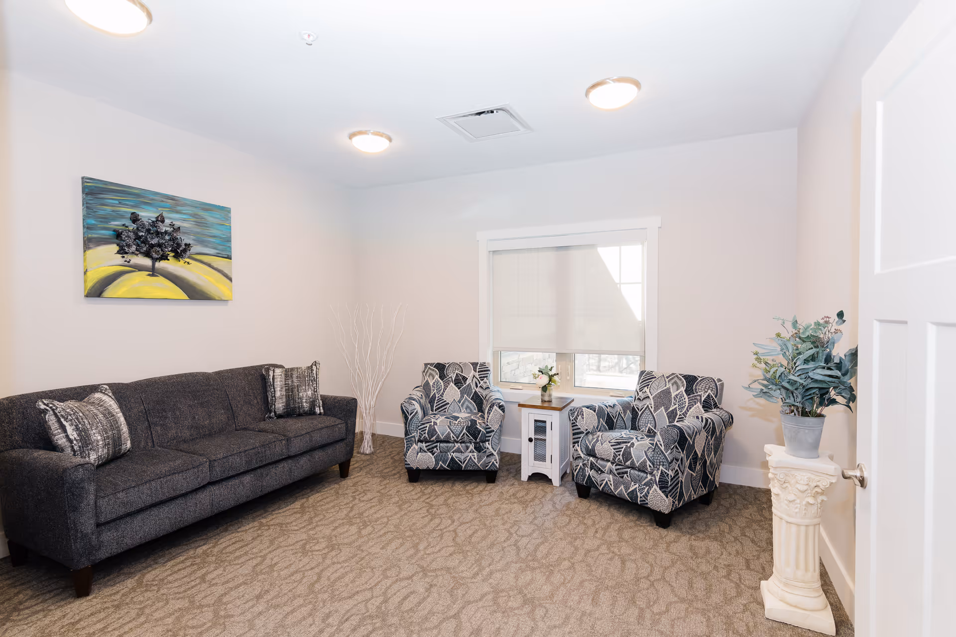 A bright sitting room with a dark gray sofa, two patterned armchairs flanking a small table under a window, wall art, and decorative plants.