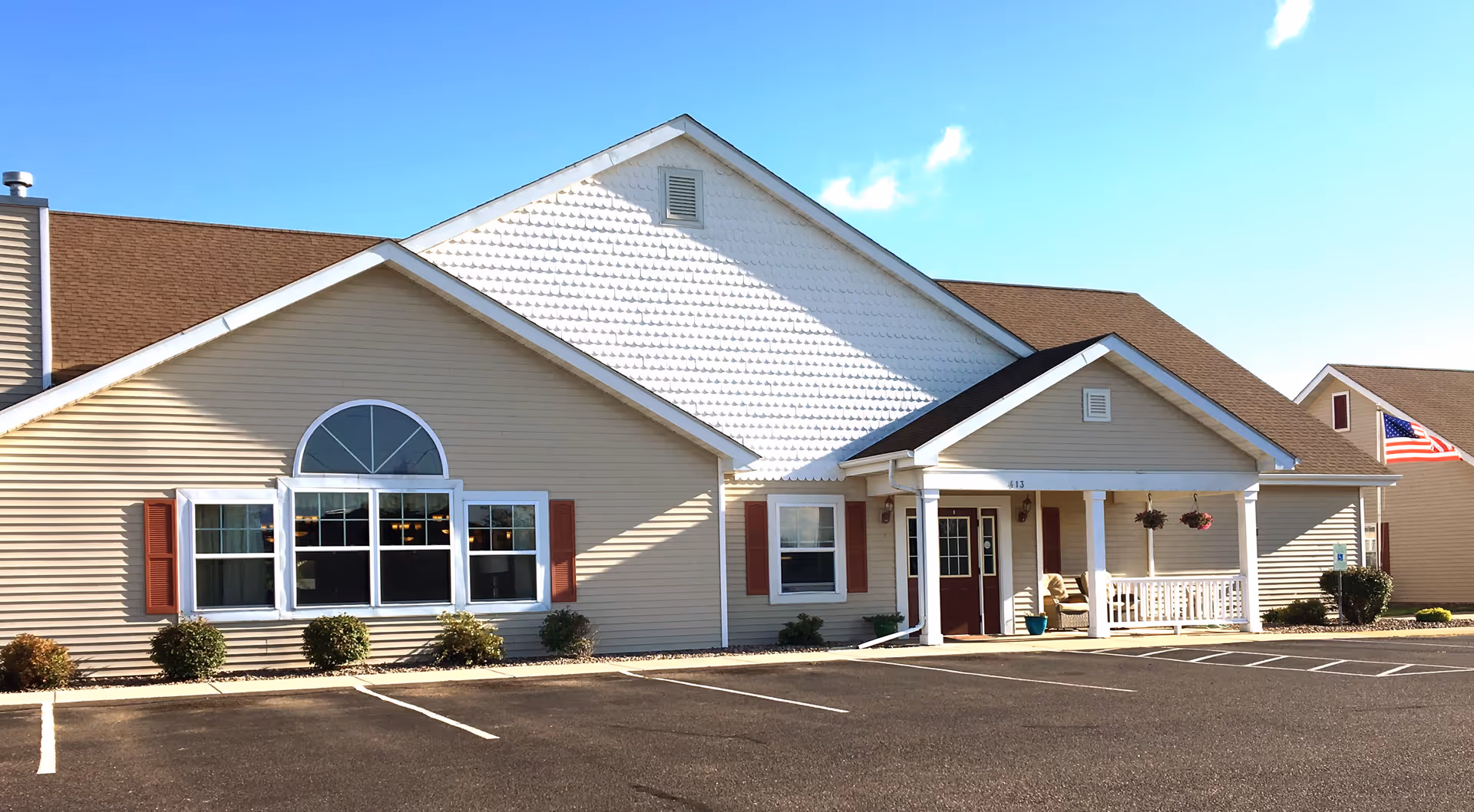Beige single-story senior living facility front with a covered entrance, large arched window, and an American flag under a clear blue sky.