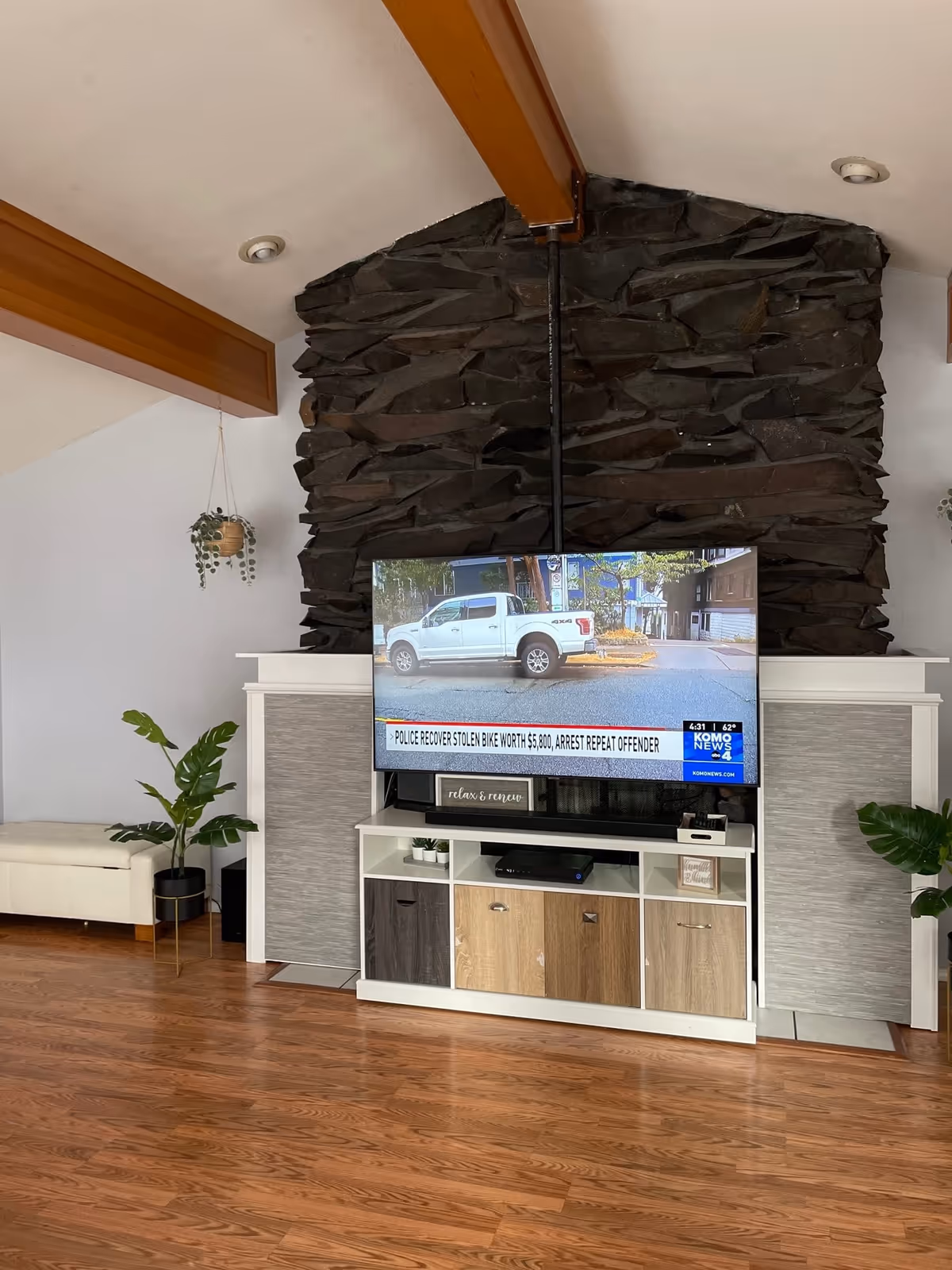 A living room area with a mounted flat-screen TV displaying a news channel. The TV is mounted on a stone fireplace with a white mantel and a white and wood-toned cabinet below it. There are two green plants in black pots on either side of the fireplace, a white bench on the left, and wooden beams on the ceiling.