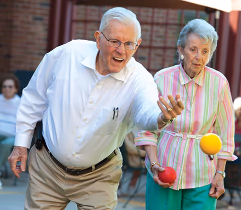 An elderly man and woman playing bocce ball outdoors. The man is in mid-throw, releasing a yellow ball, while the woman watches holding a red ball. Both are dressed casually and appear engaged in the game.