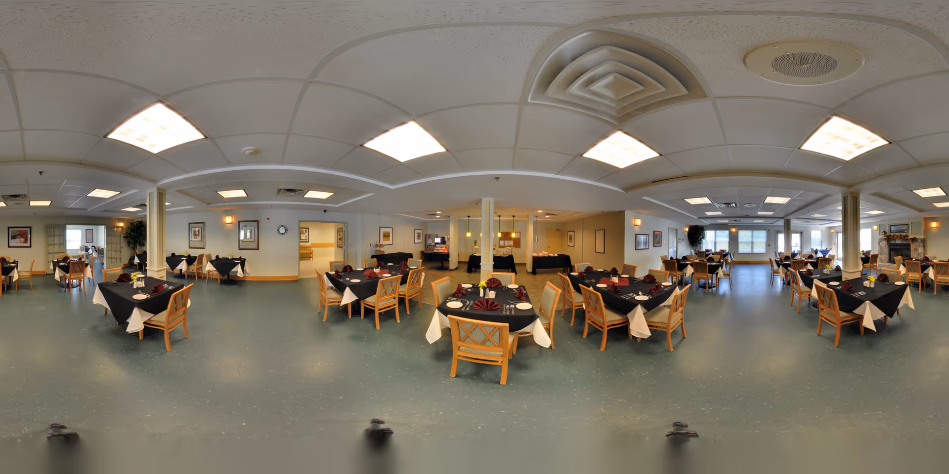A spacious dining room with multiple tables covered in black tablecloths and white napkins, each set with plates and silverware. Wooden chairs surround the tables, and the room is well-lit with ceiling lights. The walls are decorated with framed pictures, and large windows allow natural light to enter. The floor is a muted green color, and there are columns and a fireplace visible in the background.