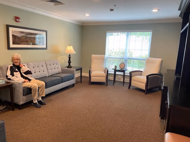 A senior woman sitting on a gray couch in a well-lit living room with beige walls. The room has two white armchairs, a small table with a clock and decorative items, a floor lamp, a large window with blinds, and a framed landscape picture on the wall.