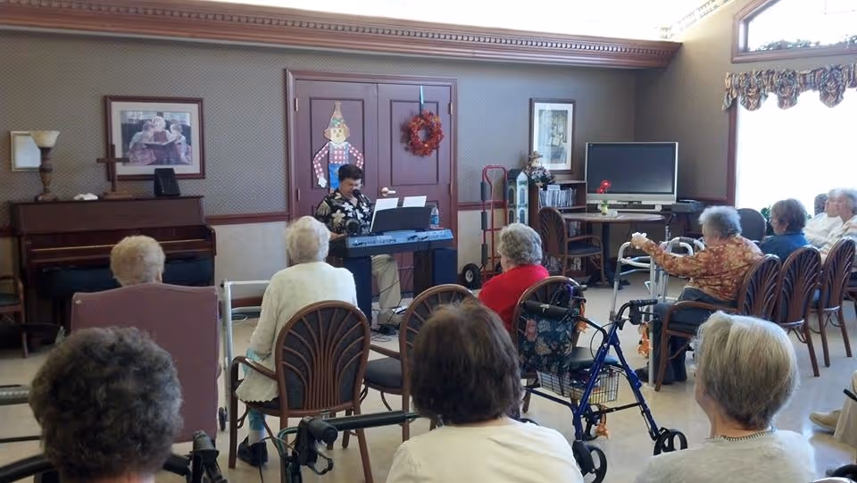 A group of elderly people seated in chairs and using walkers in a common room, attentively watching a man playing an electronic keyboard. The room has a piano, framed pictures on the walls, a television, and large windows with floral curtains letting in natural light.