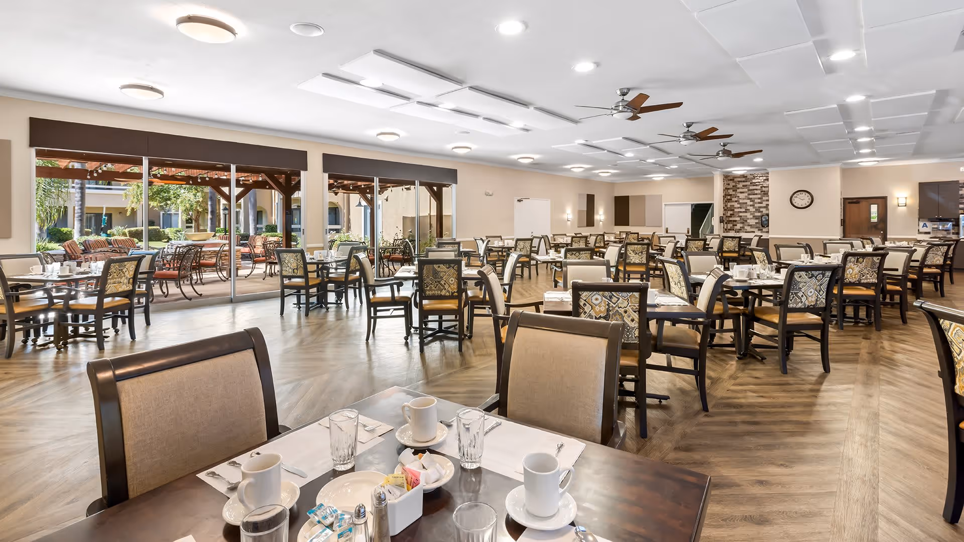 Spacious dining room with multiple tables and chairs arranged neatly, set with cups, glasses, and napkins. Large windows and sliding glass doors provide a view of an outdoor patio area with additional seating and greenery. The room features ceiling fans, recessed lighting, and wood-patterned flooring.