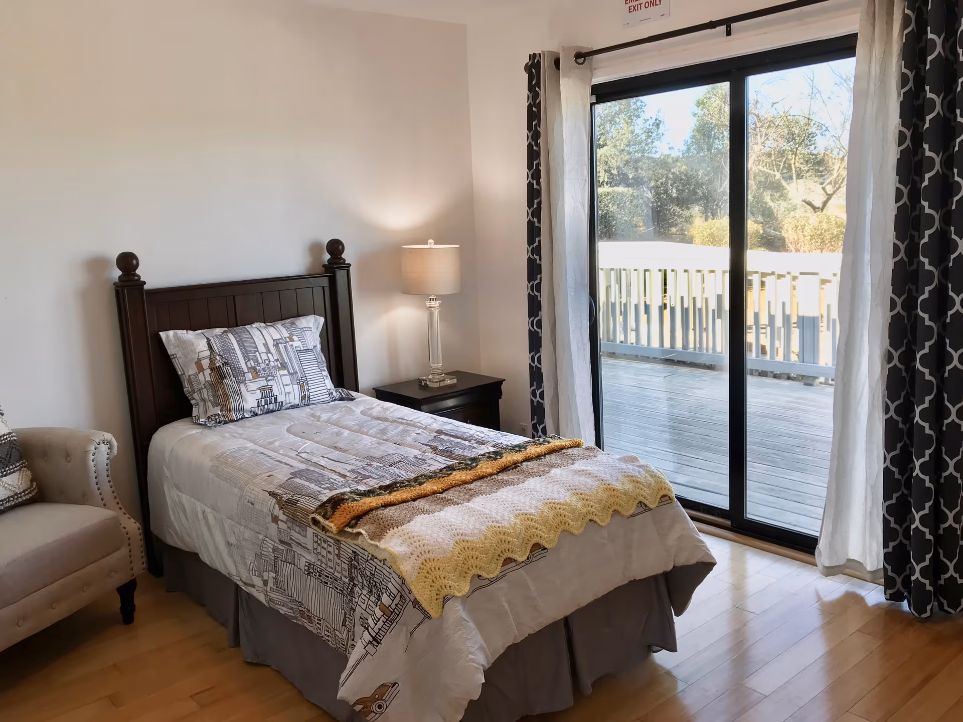A bright bedroom with a single bed, nightstand and lamp next to sliding glass doors opening onto a wooden deck.